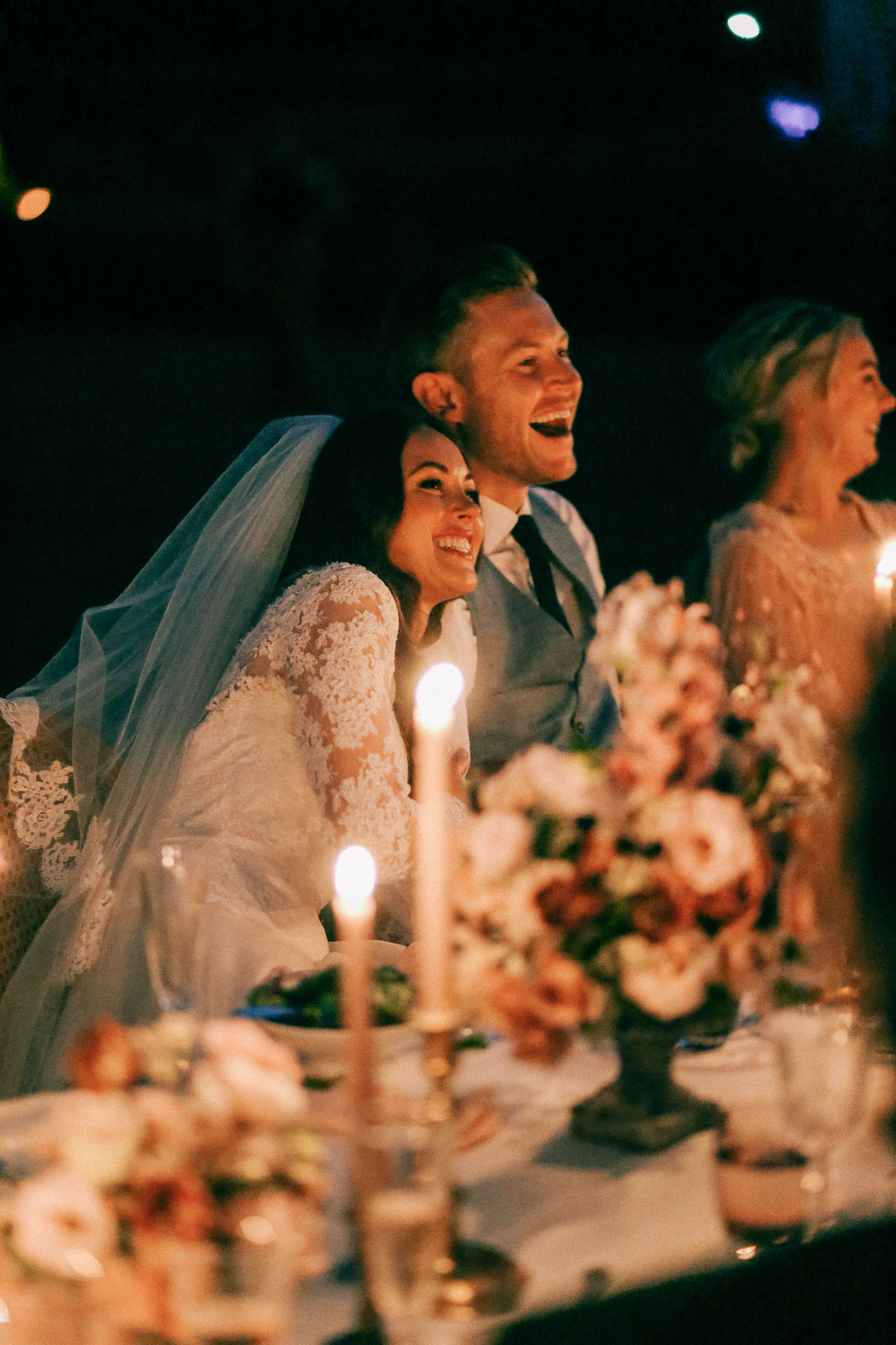 The bride and groom are seated at the head table during an evening reception, both laughing and reacting with visible joy, likely during speeches. The bride wears a long-sleeve lace gown with a long veil, and the groom is dressed in a light grey suit with a dark tie. The foreground table is decorated with lit taper candles in gold candlesticks and lush floral arrangements featuring blush, dusty rose, and terracotta blooms. A third guest, partially visible on the right and wearing a beaded or embellished blush gown, appears to be laughing alongside them. The setting is outdoors at night, with the scene lit almost entirely by warm candlelight, creating a dark, intimate atmosphere. The shot is a medium portrait taken from a low angle across the table.