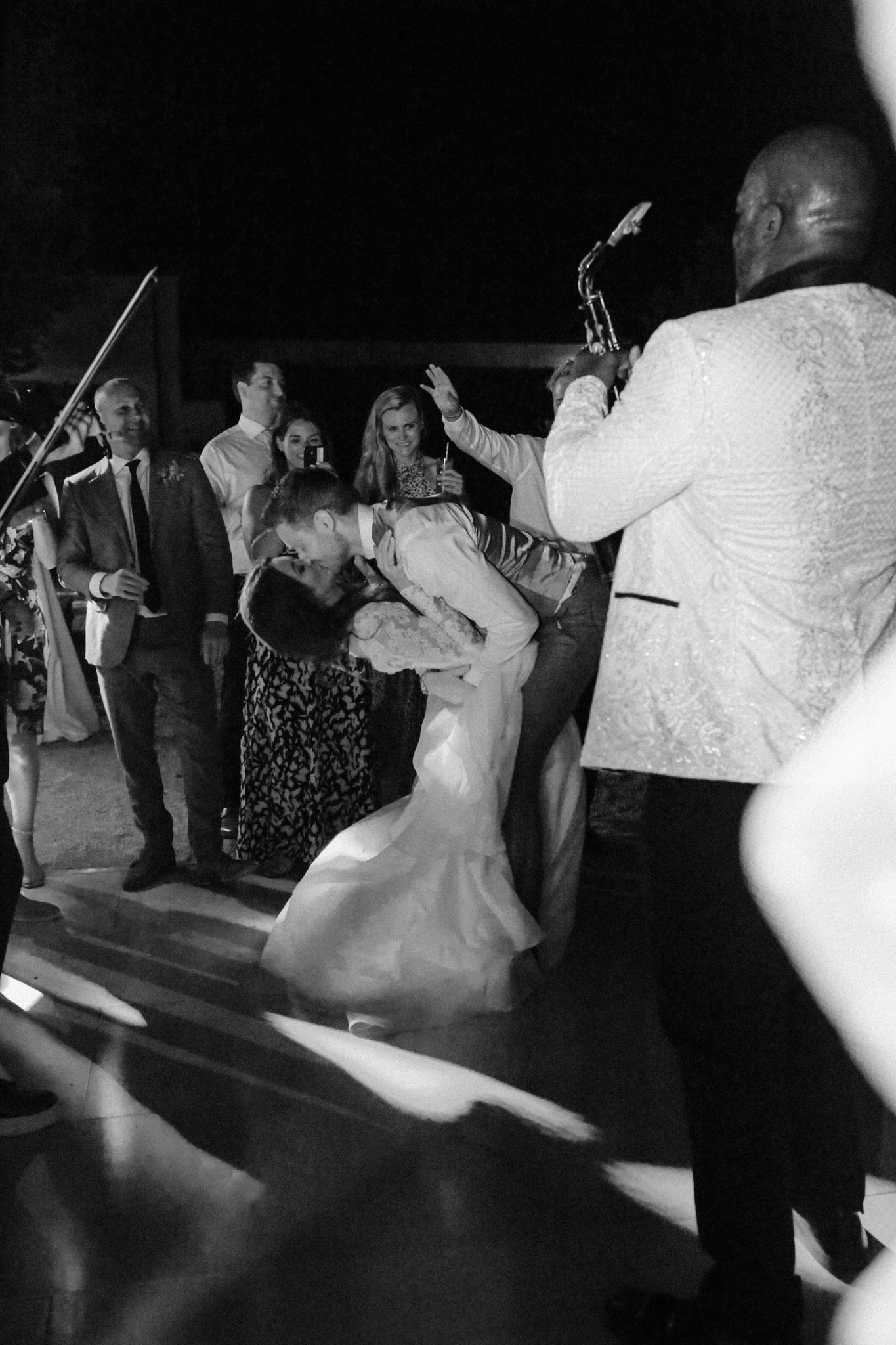 Groom dips bride for kiss on dance floor with live saxophonist performing alongside in black and white