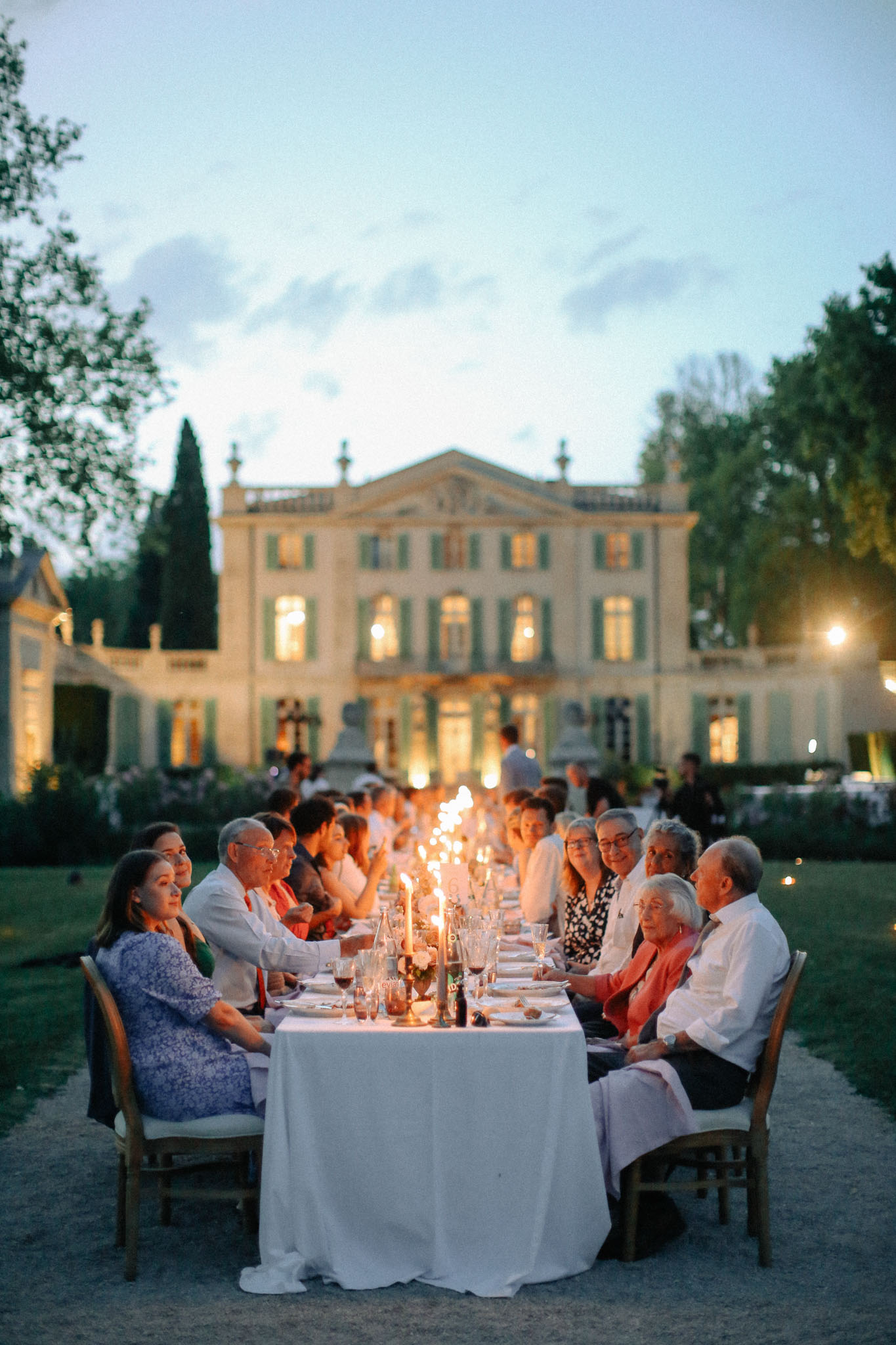 An outdoor wedding reception dinner is underway at dusk, with approximately 20–30 guests seated along a single long rectangular table covered in a white linen cloth, set on a gravel pathway in front of a French château with sage green shutters and warm interior lighting. The table is lit by tall taper candles running down its center, creating a warm glow against the blue evening light, and is set with glassware, wine bottles, and small floral arrangements. Guests are dressed in a mix of smart-casual attire including a blue floral dress, a coral blazer, and white shirts. The wide-angle shot is taken from one end of the table, looking down its length toward the illuminated château facade, which is softly lit and serves as the backdrop for the entire scene. Potential venue feature image.