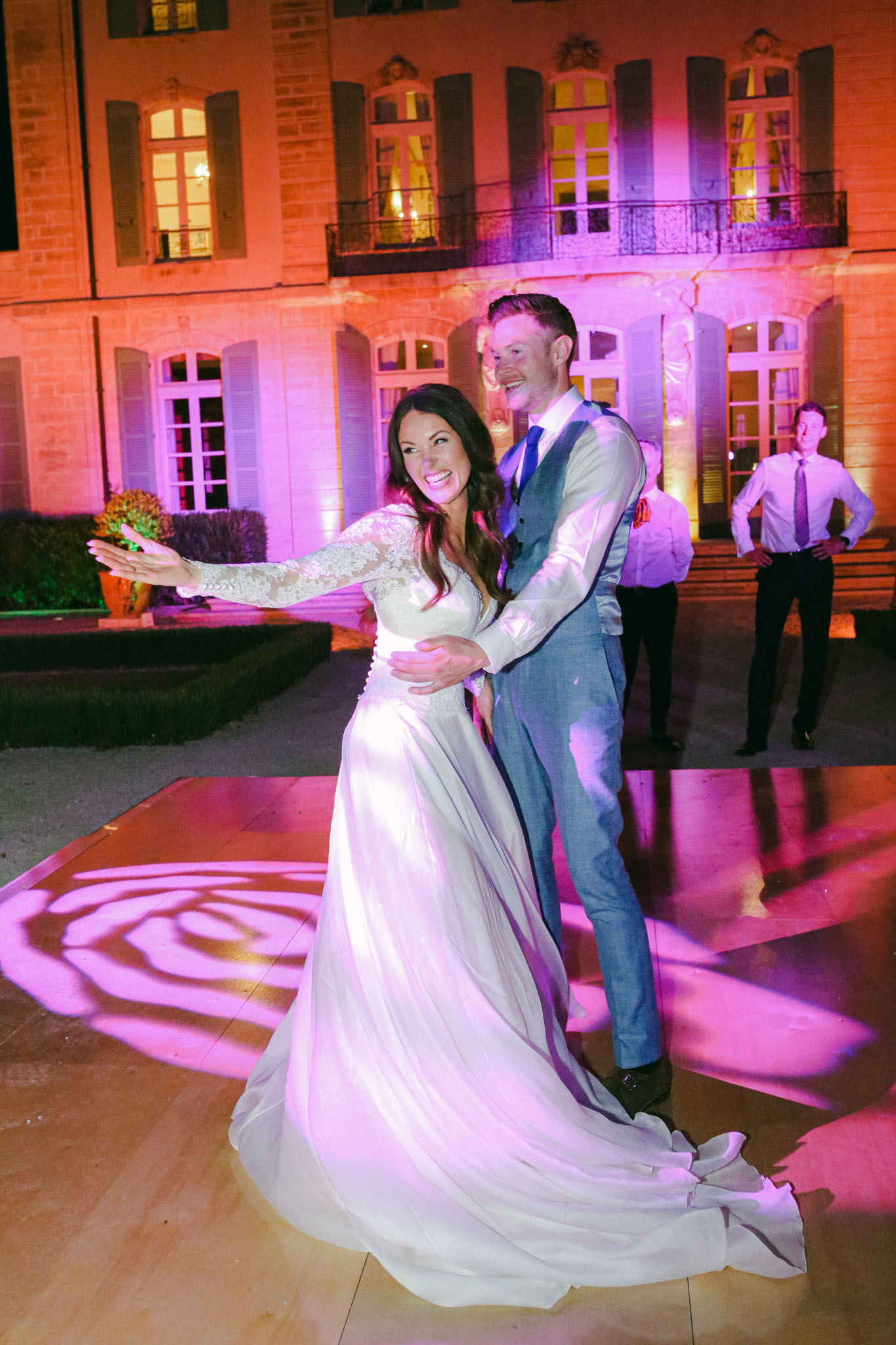 The bride and groom share their first dance on an outdoor wooden dance floor set in front of a château at night. The bride wears a white gown with long lace sleeves and a flowing skirt with a train, while the groom is dressed in a grey suit with a blue tie and waistcoat. The château facade is lit with warm orange and pink-purple uplighting, and a floral gobo pattern is projected onto the dance floor in pink-purple tones. The bride is laughing with one arm outstretched mid-spin, and the groom holds her from behind, also smiling. At least one other guest is visible in the background watching the dance. The shot is a full-length portrait taken at a slight angle, capturing the movement of the bride's skirt.