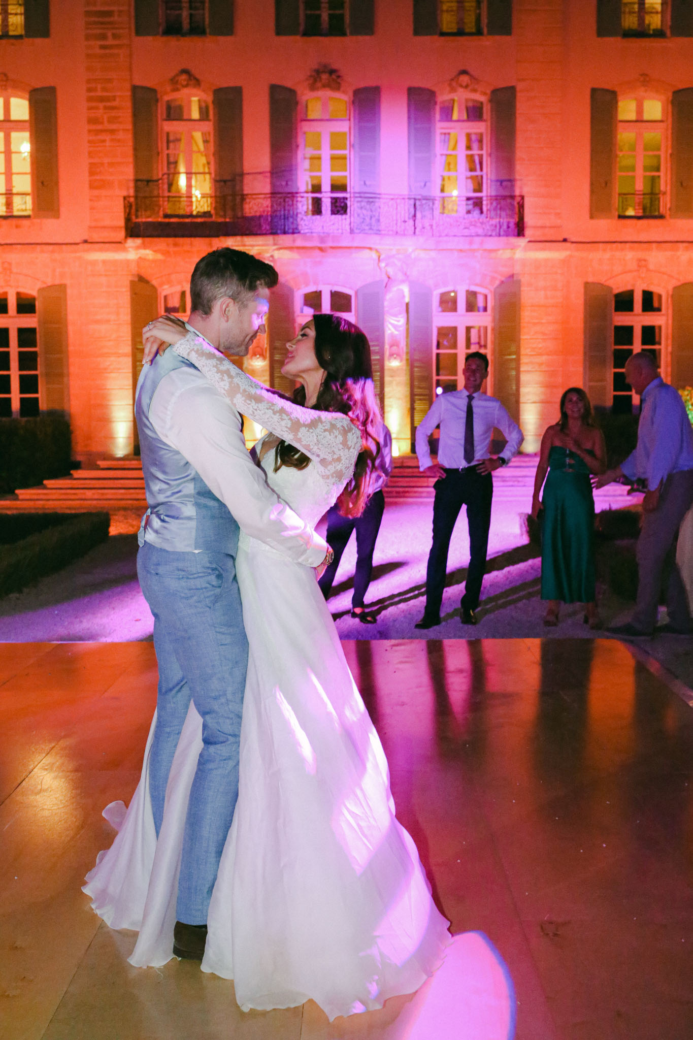 The bride and groom share their first dance on an outdoor dance floor in front of a French château at night. The bride wears a white long-sleeve lace-back gown with a flowing skirt, and the groom is dressed in a light blue suit with a matching waistcoat. The château facade is illuminated with warm orange and pink-purple uplighting, creating a dramatic evening atmosphere. A small group of approximately four guests stand in the background watching the dance, including a woman in a teal floor-length dress. The shot is a medium full-body portrait framing the couple in the foreground with the lit château architecture behind them.