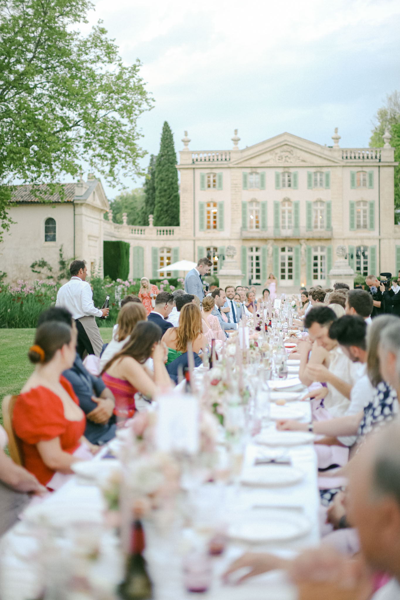 Outdoor reception dinner along long banquet table with guests and stone chateau with green shutters lit at dusk
