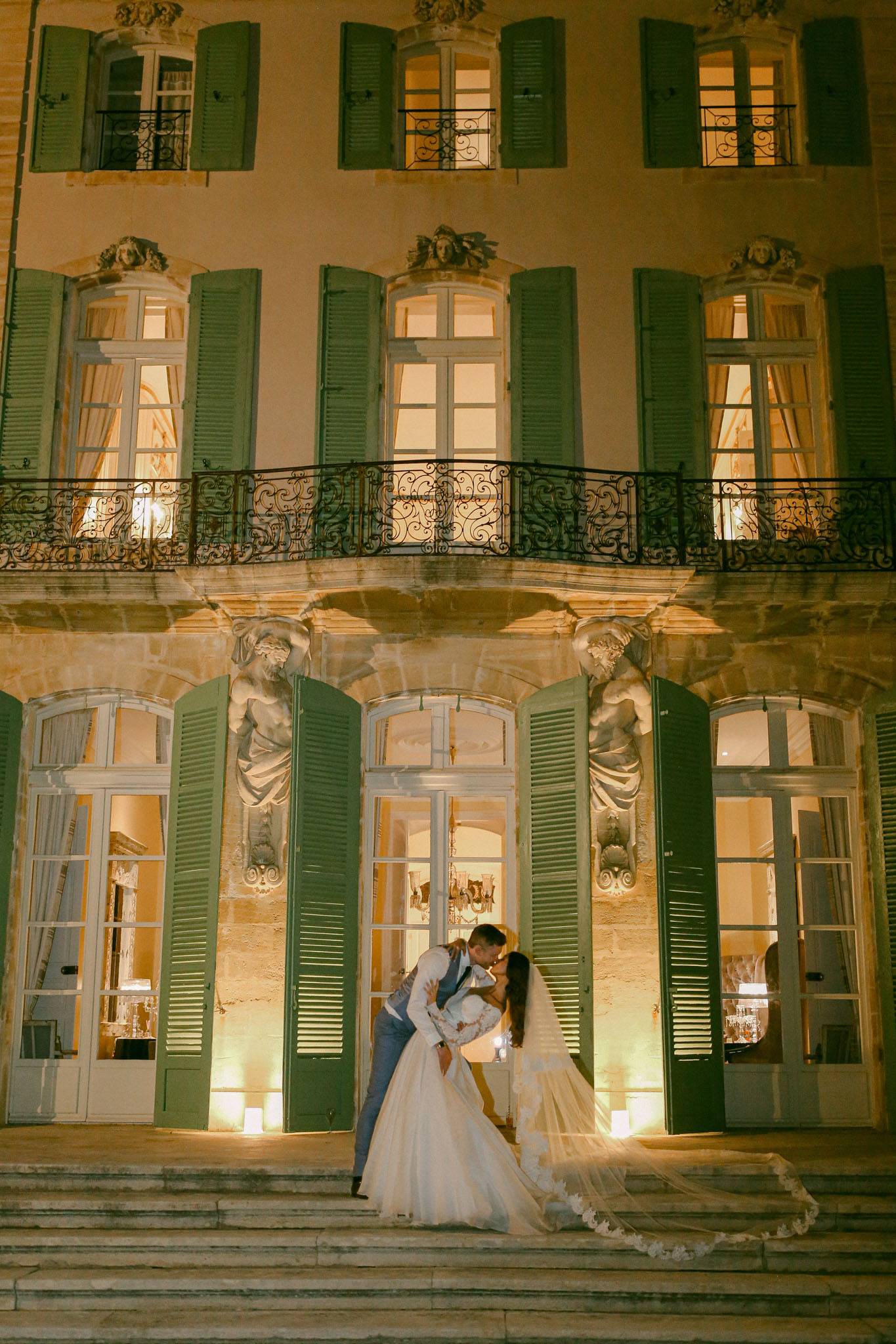 Couple sharing dip kiss on chateau steps at night with lace-trimmed veil fanned across stone stairs