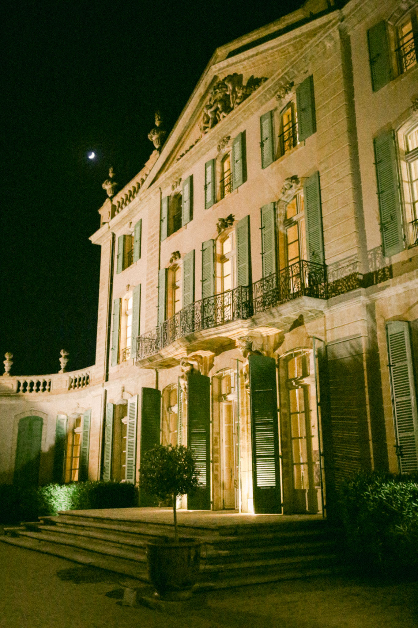 French chateau facade illuminated by warm uplighting at night with stone carvings and topiary flanking entrance
