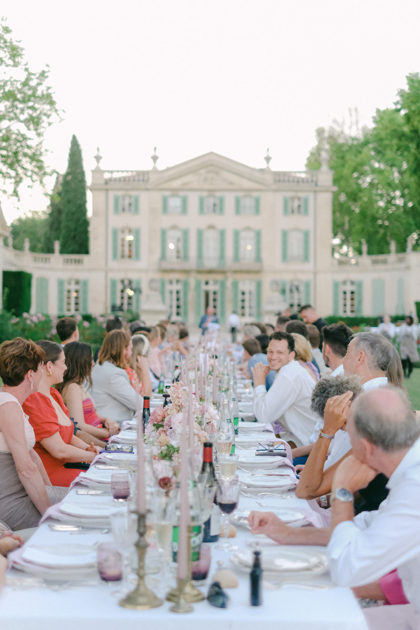 Fifty guests at long table with dusty pink rose runner and brass candlesticks before sage-shuttered chateau