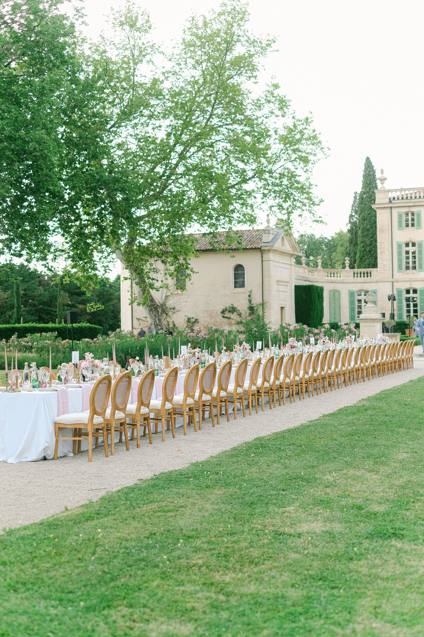 An outdoor wedding reception setup on a gravel path in the grounds of a French château, showing a very long banquet table set for a large number of guests — approximately 40 or more seats visible along one side. The table is dressed in white and soft blush pink linen with clusters of pink floral centerpieces, tall taper candles in dusty pink and nude tones, glassware, and small table number cards. Gold cane-back Louis XVI-style chairs with ivory cushioned seats are arranged along the length of the table. The pale stone château with sage green shutters, balustrades, and classical architectural detailing is visible in the background. The overall decor palette is blush pink, ivory, and gold, with a classic French formal styling. Wide shot taken from a low angle along the side of the table, emphasizing the scale of the setup. Potential venue feature image.