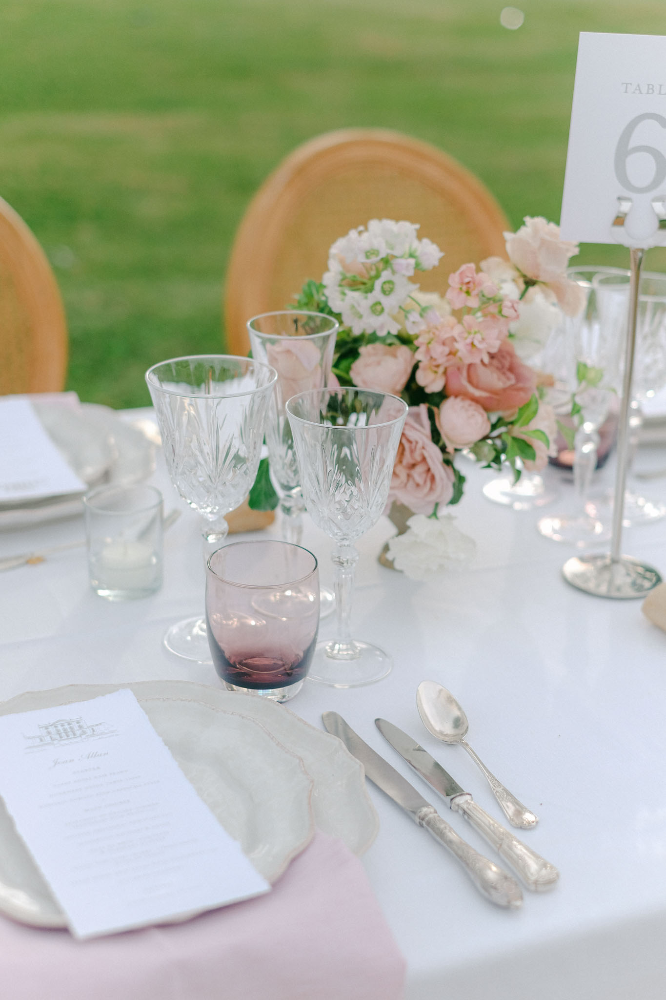 Close-up detail shot of an outdoor wedding reception table setting, likely table number 6 as indicated by a white card in a silver holder. The white linen tablecloth is set with silver-edged charger plates, a linen napkin in a neutral grey-beige tone, silver vintage-style cutlery including two knives and a spoon, cut-crystal wine glasses, and a smoky mauve-tinted lowball glass. A printed menu card with an illustrated venue header is placed at the setting. The centerpiece features blush pink garden roses, soft white blooms, and green foliage. Natural wood bistro-style chairs are visible in the background. The overall decor palette is blush pink, white, and silver with a classic French garden aesthetic.