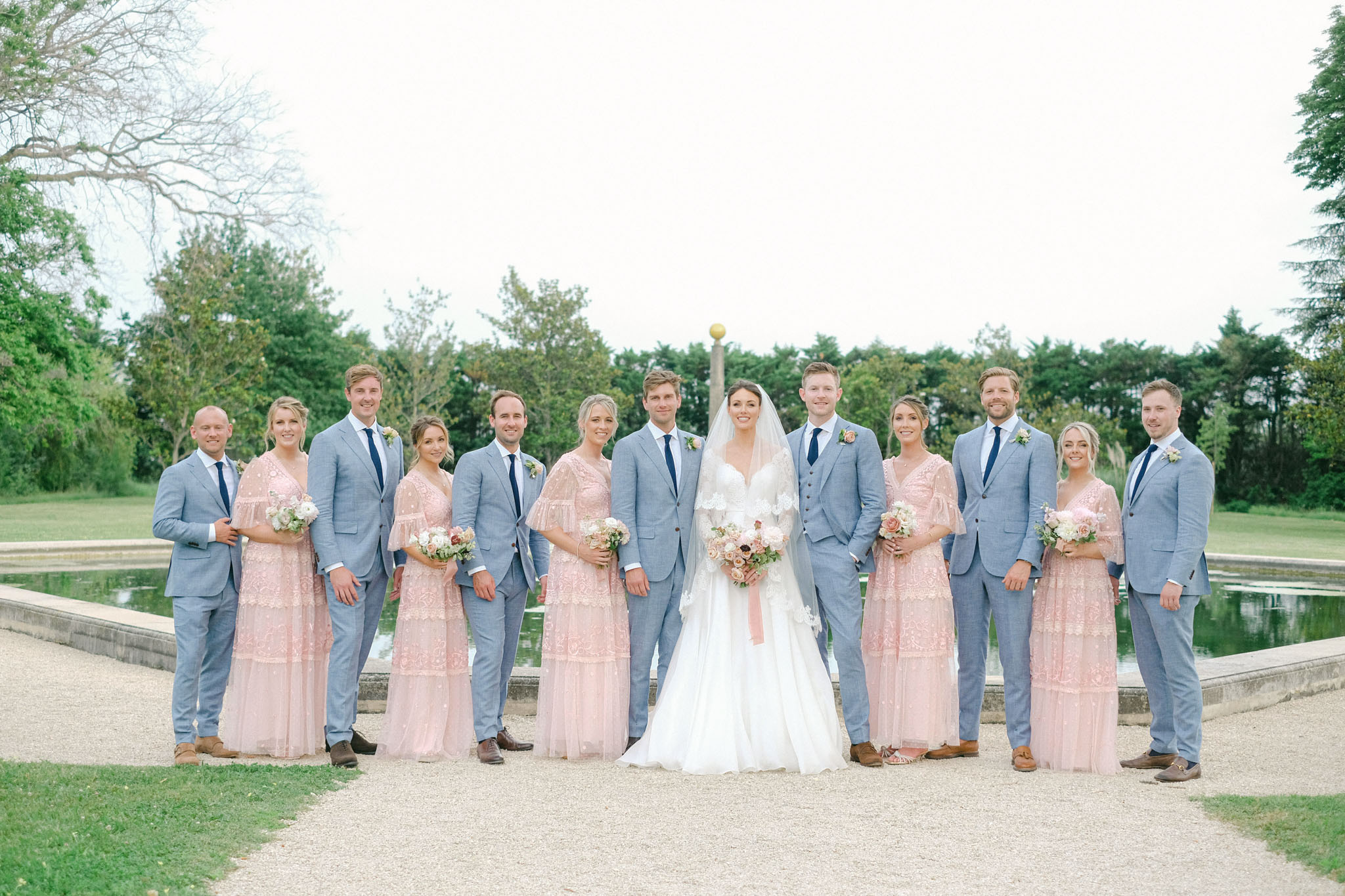 A full bridal party portrait taken outdoors on a gravel path beside a rectangular ornamental pool, set within formal garden grounds. The group of twelve — the bride, groom, four bridesmaids, and five groomsmen — stands in a single line facing the camera. The bride wears a white A-line gown with lace long sleeves and a cathedral-length veil, holding a bouquet of blush, ivory, and dusty rose blooms. The bridesmaids wear floor-length blush pink tiered lace dresses with flutter sleeves, each carrying small rounded bouquets of blush and ivory flowers. The groomsmen are dressed in light chambray blue suits with navy ties and small floral buttonholes. The overall color palette is soft blush, chambray blue, and ivory, giving the styling a classic yet light, airy feel. The composition is a wide group shot with the couple positioned at the center.
