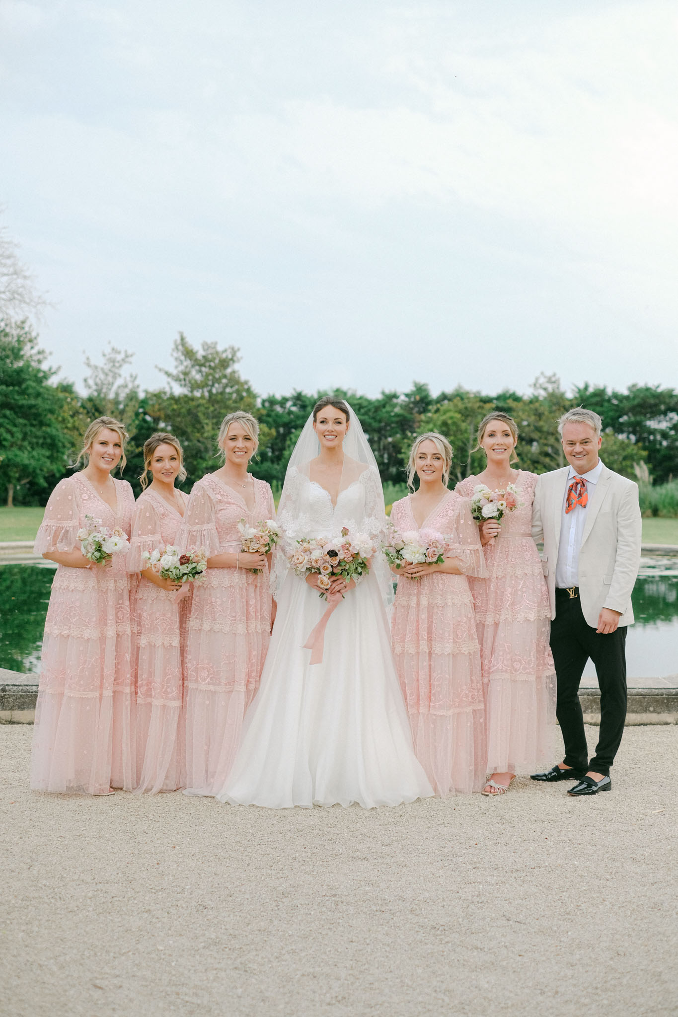 A bridal party portrait taken outdoors on a gravel path in front of a ornamental pond or reflecting pool. The bride stands at center in a white ballgown with a sweetheart neckline and a long cathedral-length veil, holding a loose bouquet of blush, ivory, and peach roses and anemones with a blush ribbon trailing. She is flanked by five bridesmaids wearing matching blush pink tiered lace midi-to-maxi dresses with flutter sleeves and V-necklines, each holding smaller versions of the bridal bouquet with ivory and blush blooms. One male attendant stands at far right in a cream blazer, black trousers, patent loafers, and a patterned orange and red tie. The group is composed in a wide portrait shot with all seven subjects facing the camera and smiling.
