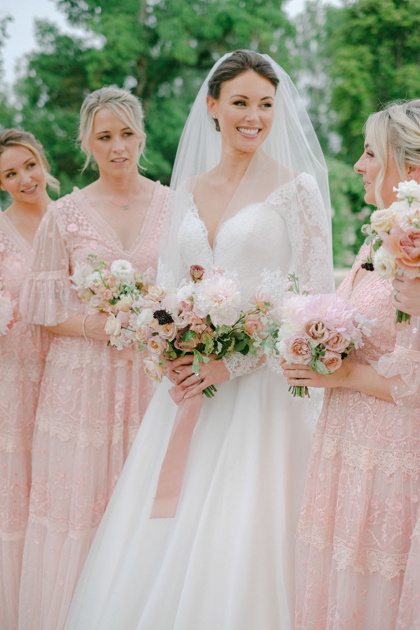 Bride in white lace gown with three bridesmaids in mismatched blush lace dresses holding peony bouquets