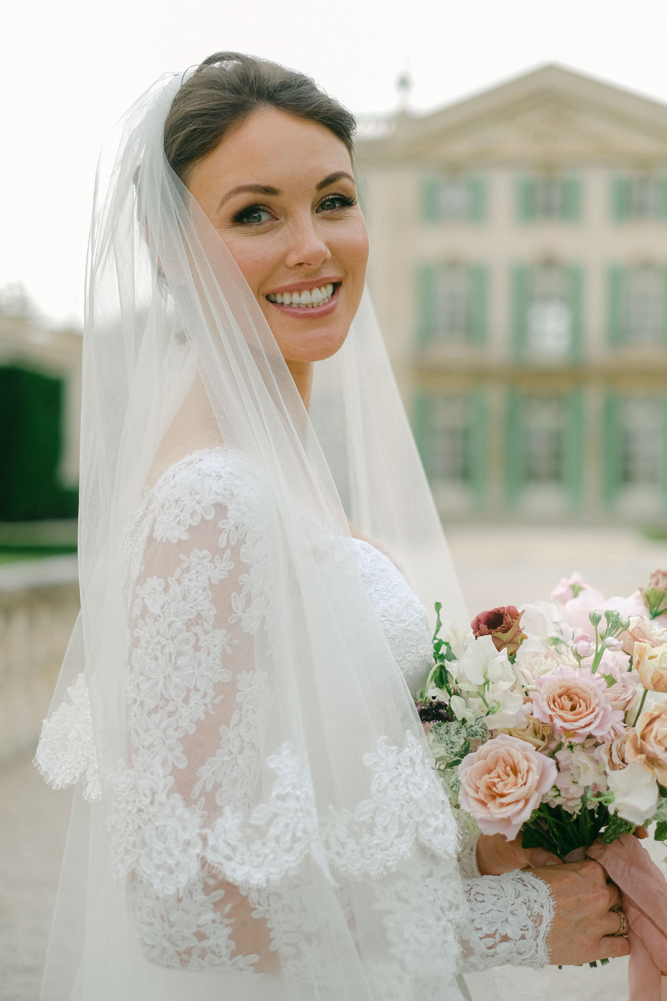 Bride looking over shoulder holding blush and peach rose bouquet with chateau in background