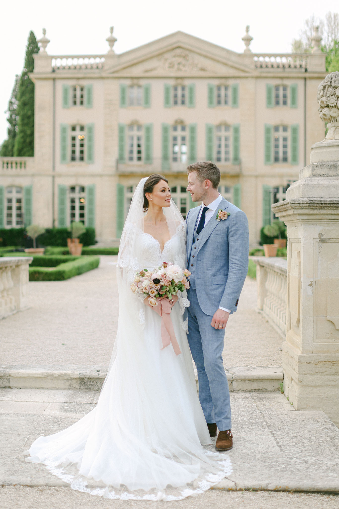 A couple portrait taken outdoors on the stone terrace of a French château, with the main building visible and softly blurred in the background. The bride wears a white A-line gown with a lace-trimmed bodice, long lace sleeves, and a cathedral-length veil with a lace hem; she holds a loose bouquet of blush, dusty rose, and burgundy garden roses with trailing dusty pink ribbon. The groom wears a light blue three-piece suit with a navy tie and a blush rose boutonnière; the two are turned toward each other, with the groom looking at the bride. The château behind them features pale stone architecture with green shutters, a formal parterre garden with clipped hedges, and decorative stone balustrades, lending a classic French estate setting to the image. Potential venue feature image.
