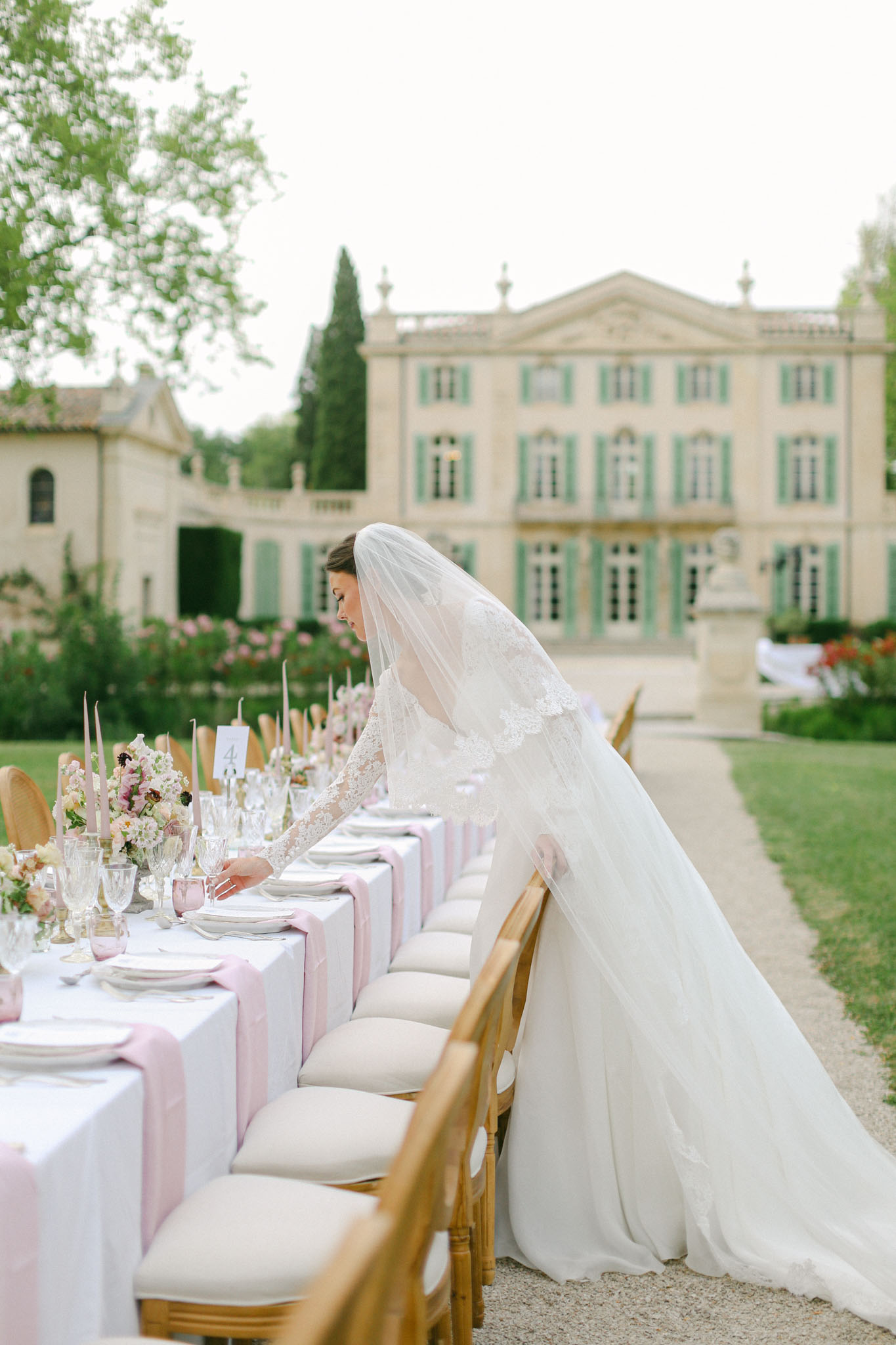 Bride in lace-sleeve ballgown adjusts blush and mauve reception table before chateau with sage shutters
