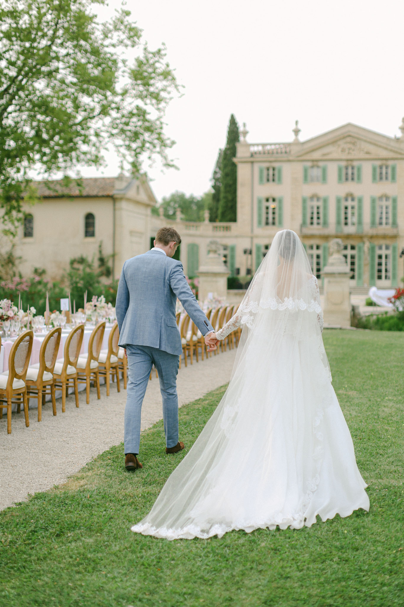 The bride and groom walk hand-in-hand toward a French château with pale stone façade and sage green shutters, photographed from behind in a wide portrait shot. The bride wears a full-length white ball gown with lace long sleeves and a cathedral-length veil trimmed with lace appliqué detailing; the groom wears a light blue suit with brown leather shoes. To their left, a long outdoor reception table is set on a gravel path, lined with natural wood and cane-back chairs, tall taper candles, and arrangements of what appear to be blush pink florals, styled in a classic French garden aesthetic. Potential venue feature image.