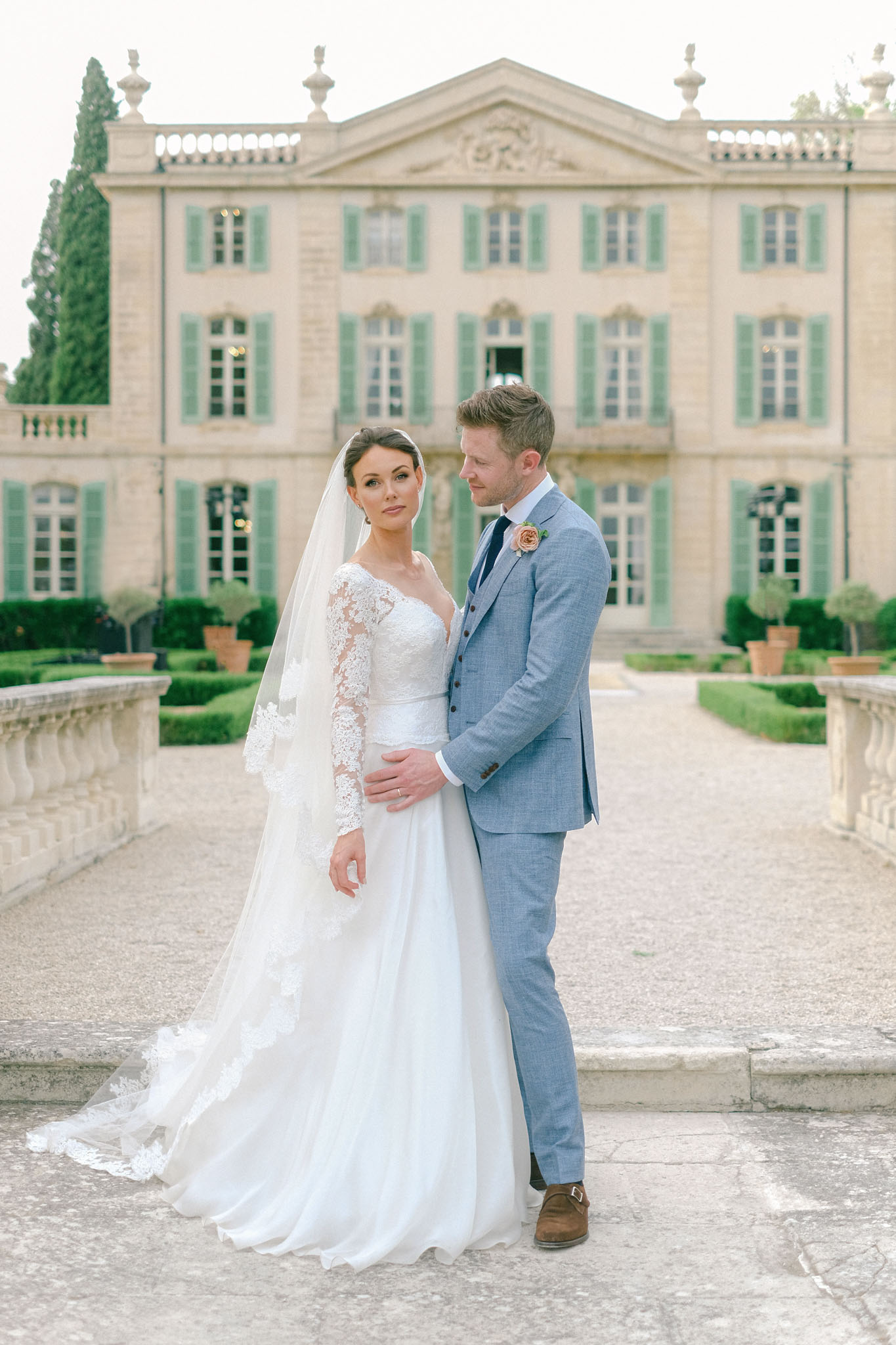 A couple portrait taken outdoors on the forecourt of a French château, with the main façade softly out of focus behind them. The bride wears a white A-line gown with long lace illusion sleeves, a deep V-neckline, and a lace-edged cathedral veil; her dark hair is pulled back in an updo. The groom wears a light blue three-piece suit with a navy tie and a single blush pink rose boutonnière, paired with tan suede monk-strap shoes. He stands behind her with one hand at her waist, looking down at her, while she faces the camera directly. The château behind them features pale stone architecture with sage green shutters, ornate balustrades, and formally clipped boxwood hedging flanking a central path, suggesting a classic French estate setting. The overall styling is classic and polished. Potential venue feature image.