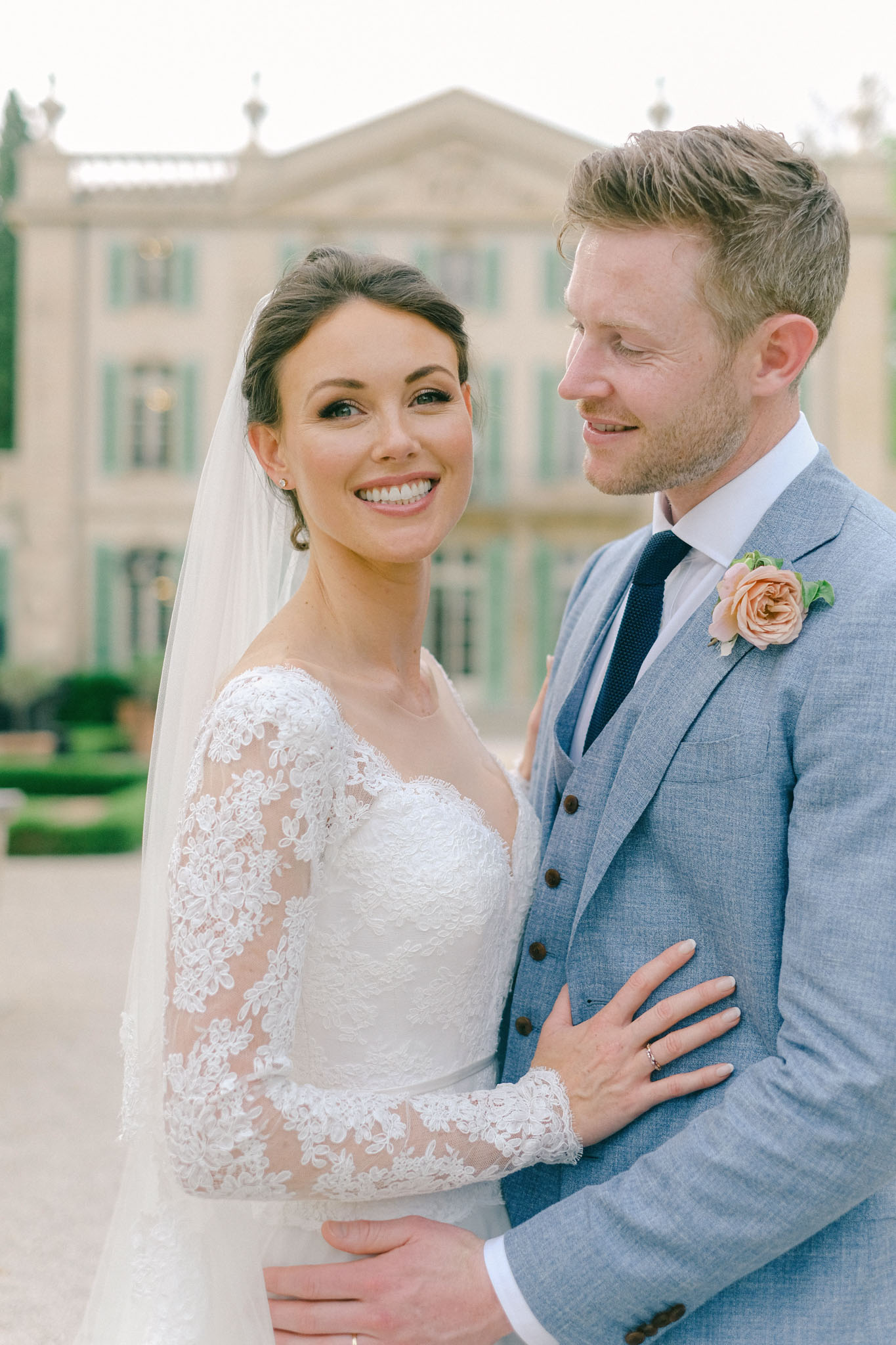 Bride in long-sleeve lace gown with cathedral veil and groom in blue suit smiling at chateau gardens