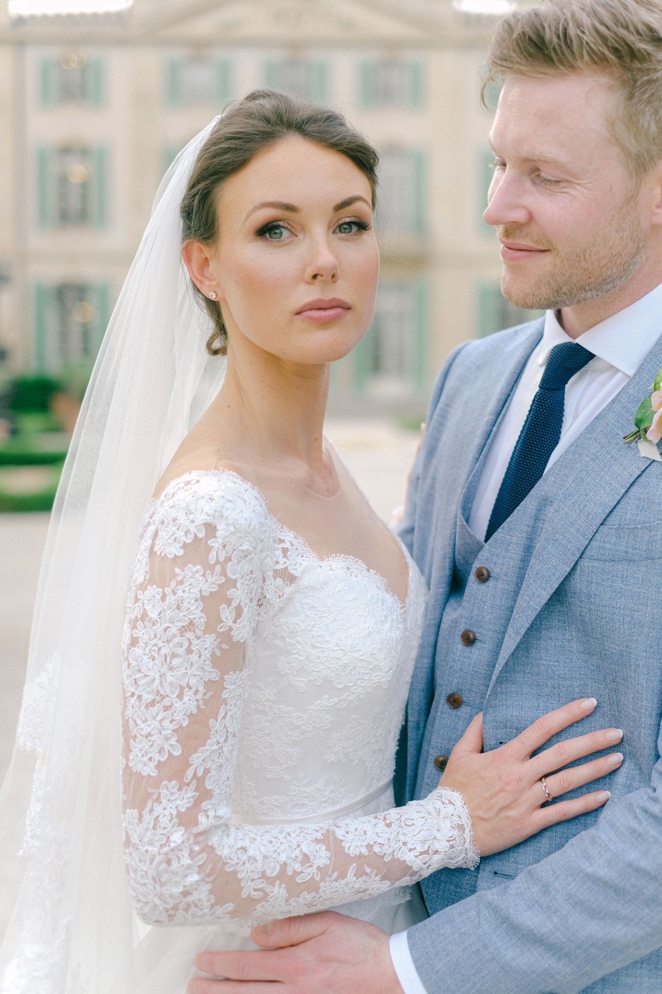 A close-up couple portrait taken outdoors in front of a blurred French château facade with pale stone architecture and sage-green shuttered windows visible in the background. The bride wears a white lace wedding dress with long sheer lace sleeves, a sweetheart neckline, and a cathedral-length veil with her dark hair swept up; she looks directly into the camera. The groom wears a light blue three-piece suit with a navy knit tie and a blush floral boutonnière, and he looks downward with a slight smile. The bride's left hand, showing a diamond ring, rests on the groom's arm. The image is a tight portrait composition with soft natural light and a shallow depth of field.