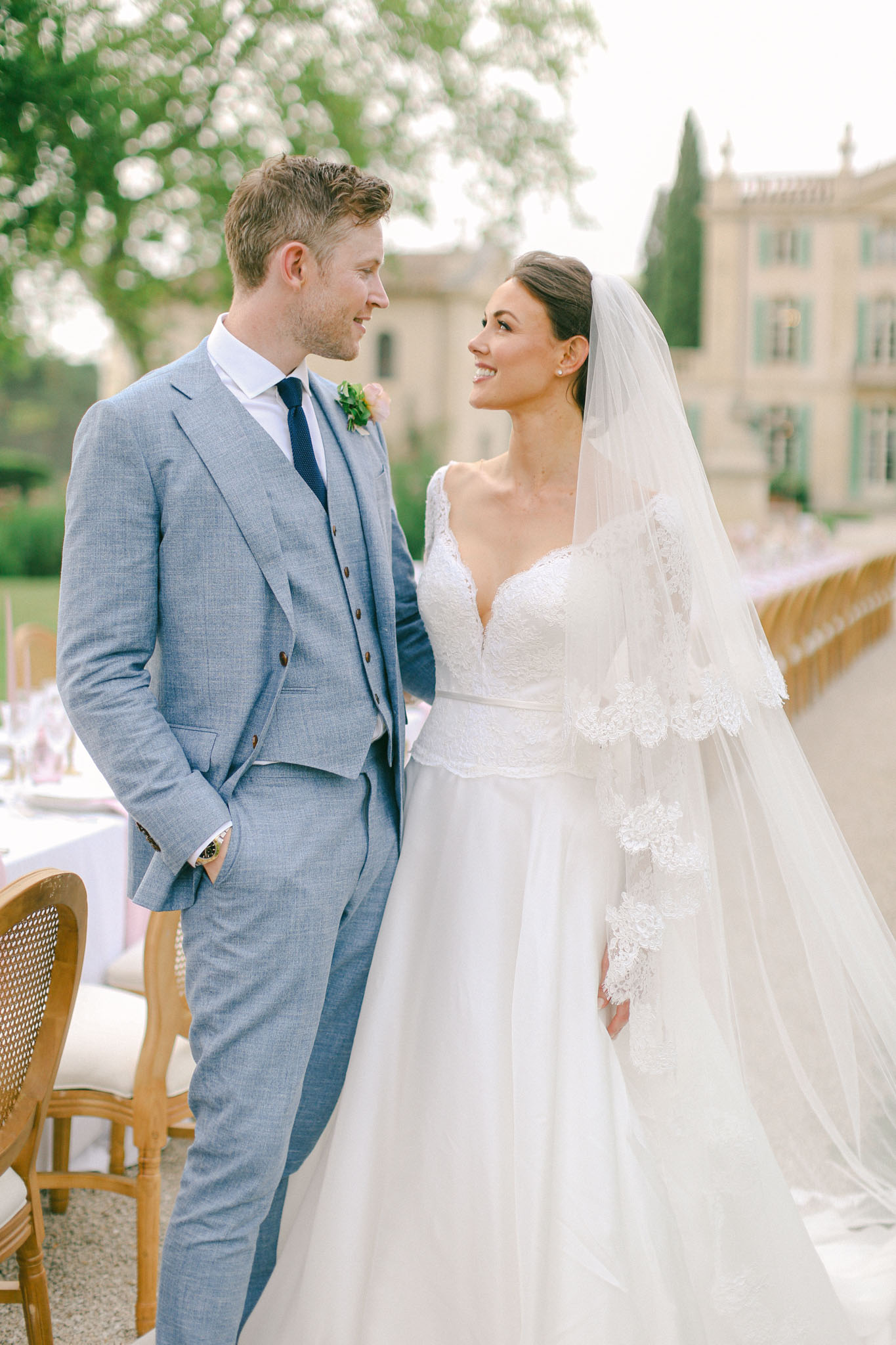 A couple portrait taken outdoors on a terrace at what appears to be a French château, visible in the background with pale stone architecture and green shutters. The groom wears a light blue three-piece suit with a navy tie and a small greenery boutonniere, while the bride wears a white ball gown with long lace appliqué sleeves, a deep V-neckline, and a long cathedral-length veil with lace trim. The bride has her dark hair in an updo and is smiling at the groom, who looks back at her. In the background, a long outdoor reception table dressed in white linens with pale pink accents and wooden bistro-style chairs is partially visible, suggesting a classic French garden reception setup. The image is a mid-length portrait with soft, bright natural lighting.