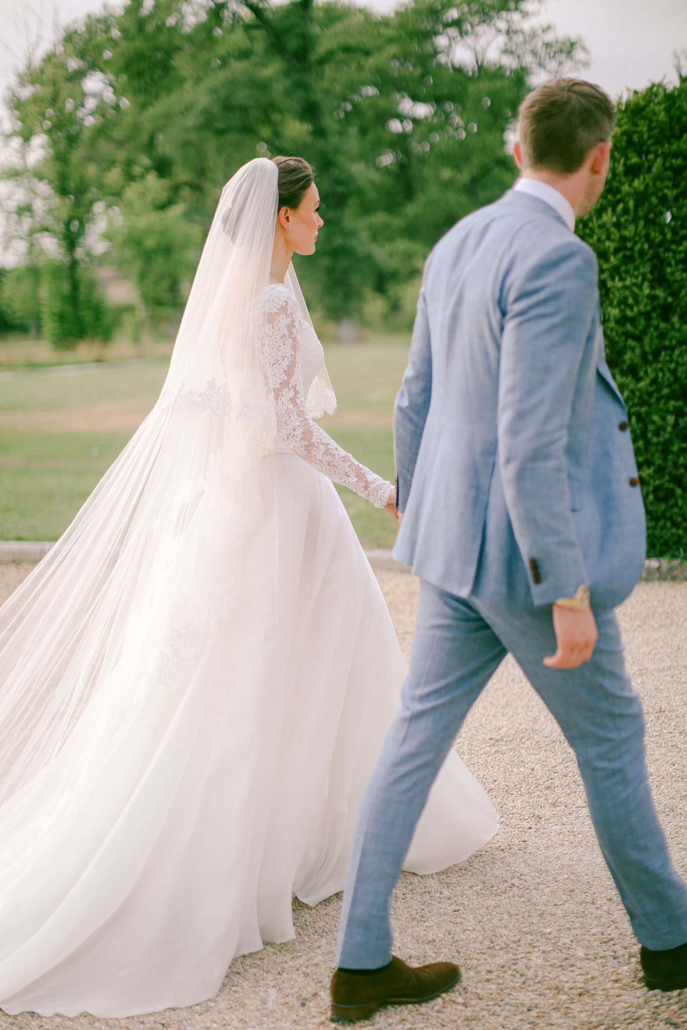 Bride in ivory lace gown with cathedral veil and groom in light blue suit walking from behind on gravel path
