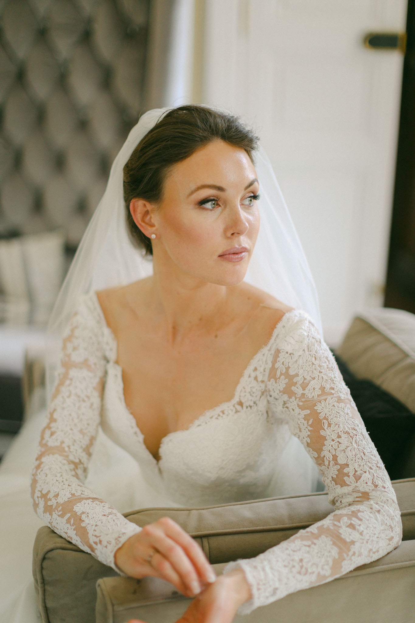 Bride in ivory lace gown with sheer sleeves and cathedral veil seated on grey sofa during getting ready