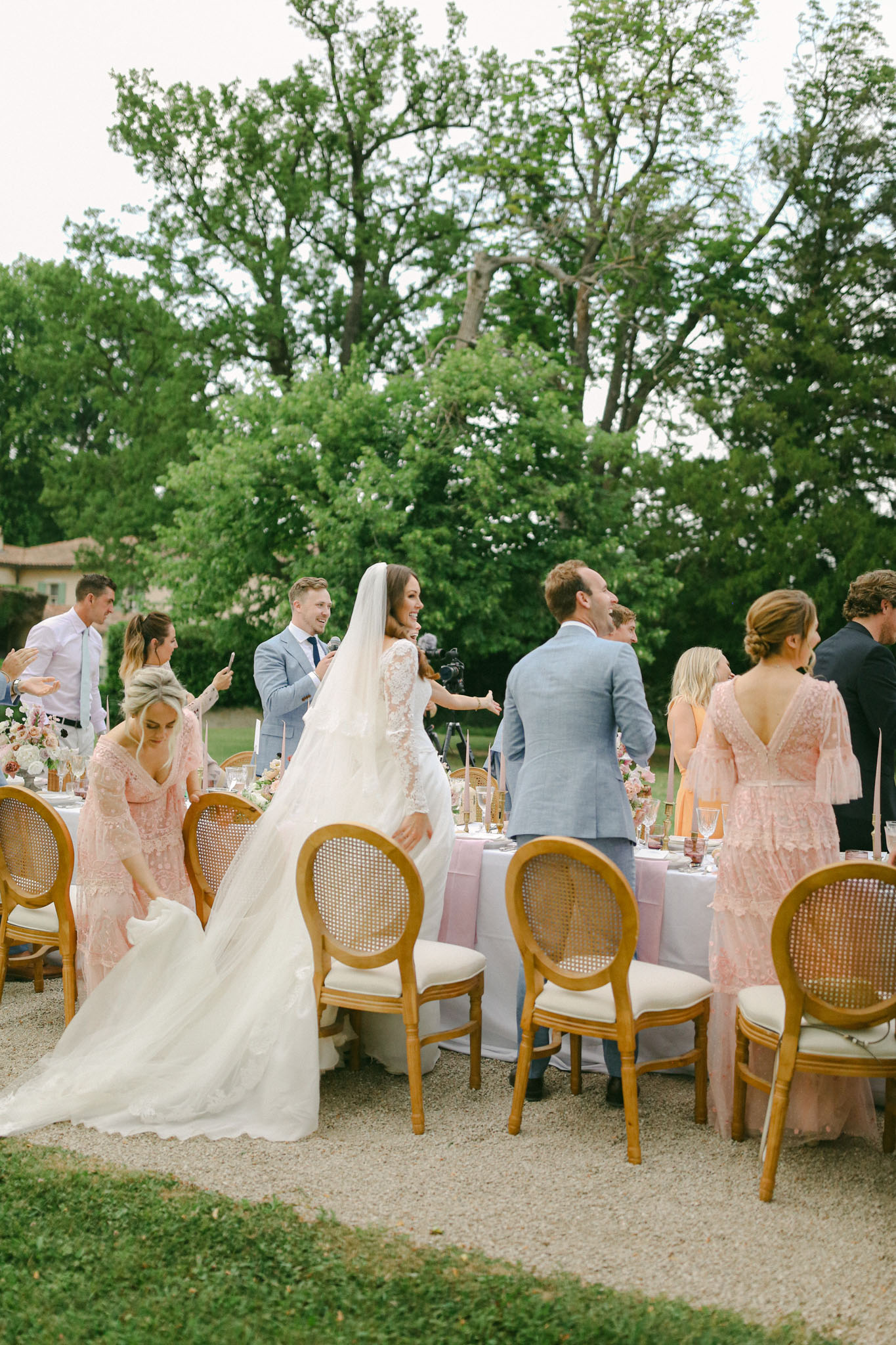 An outdoor wedding reception speech moment captured in a wide shot, set on a gravel terrace surrounded by mature trees, with a château building partially visible in the background. The groom, wearing a light blue suit, holds a microphone and speaks while the bride stands beside him in a long-sleeve lace wedding gown with a cathedral-length veil and an extended train being held by a bridesmaid. Approximately ten guests and bridal party members are gathered around a long rectangular dining table covered in a dusty lavender linen, set with gold candlestick holders, floral centerpieces featuring blush and coral blooms, glassware, and pink napkins. The bridesmaids are dressed in blush pink lace midi dresses with flutter or long sleeves, and the male guests wear suits in light blue and navy. Seating consists of natural wood round-back cane chairs with cream cushions. The overall styling is romantic and garden-party in feel, with a soft blush, lavender, and gold color palette.