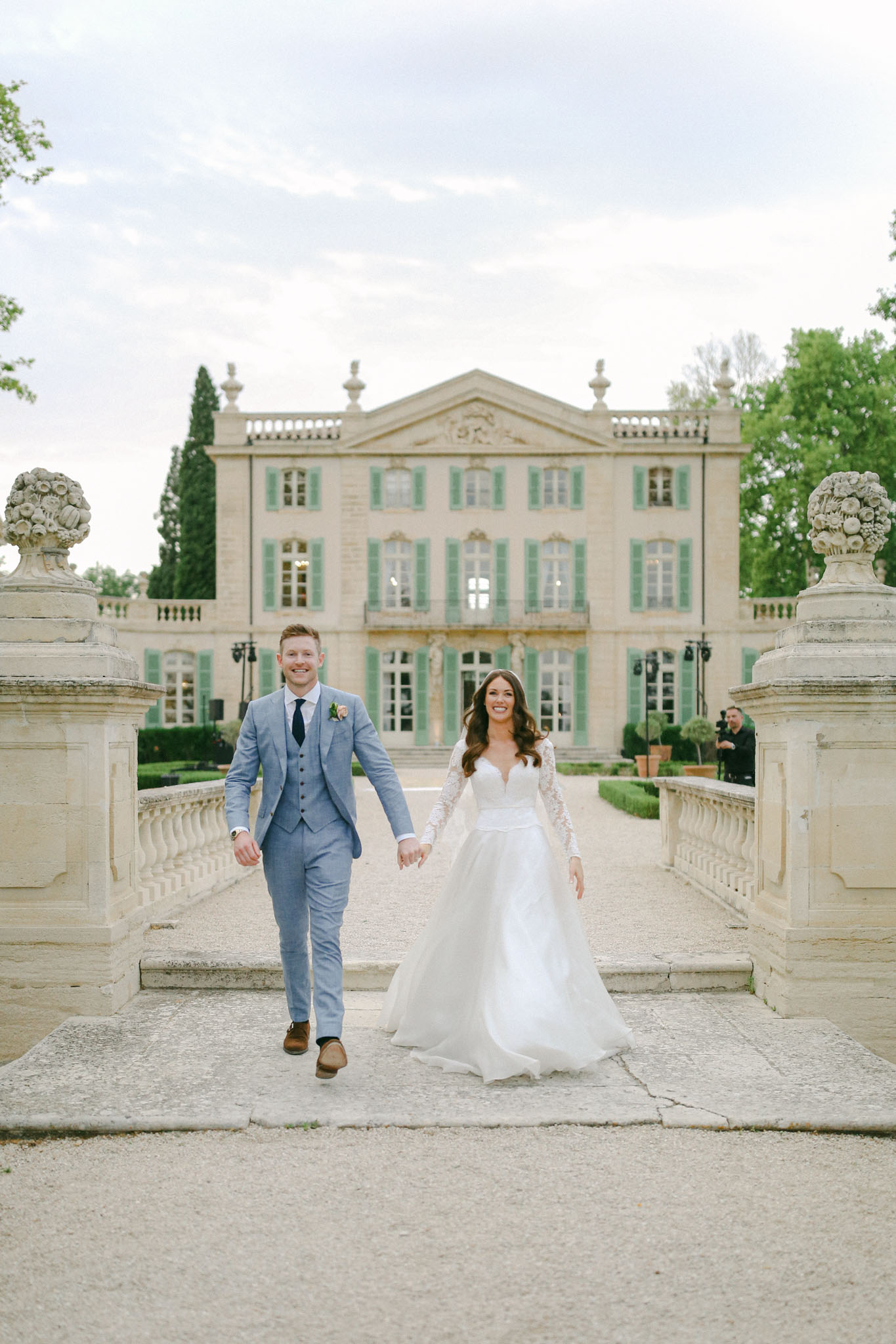 Bride and groom walking on chateau forecourt with green shutters topiary and stone balustrades