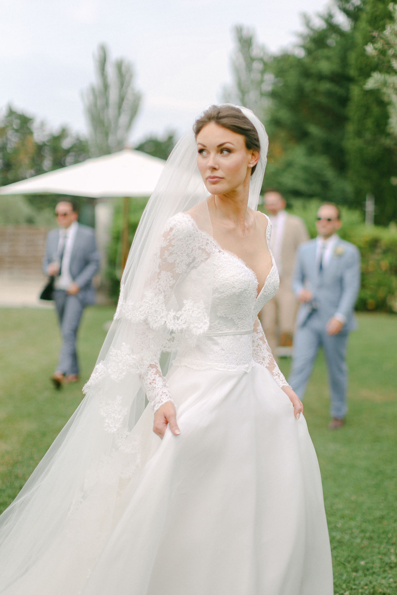 Bride in white V-neck gown with lace sleeves and cathedral veil on manicured lawn with guests beyond