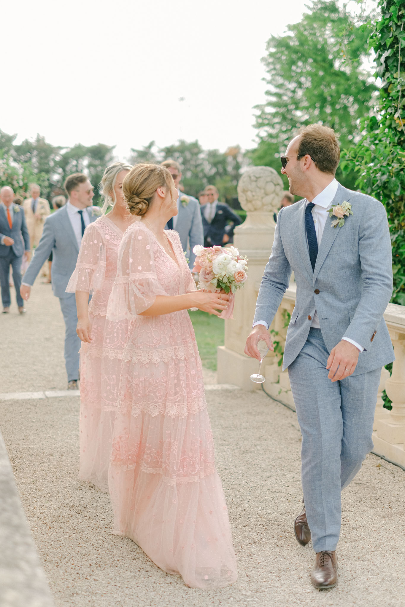 Bridesmaid in blush tiered lace dress and groomsman in grey suit walking with champagne during cocktail hour