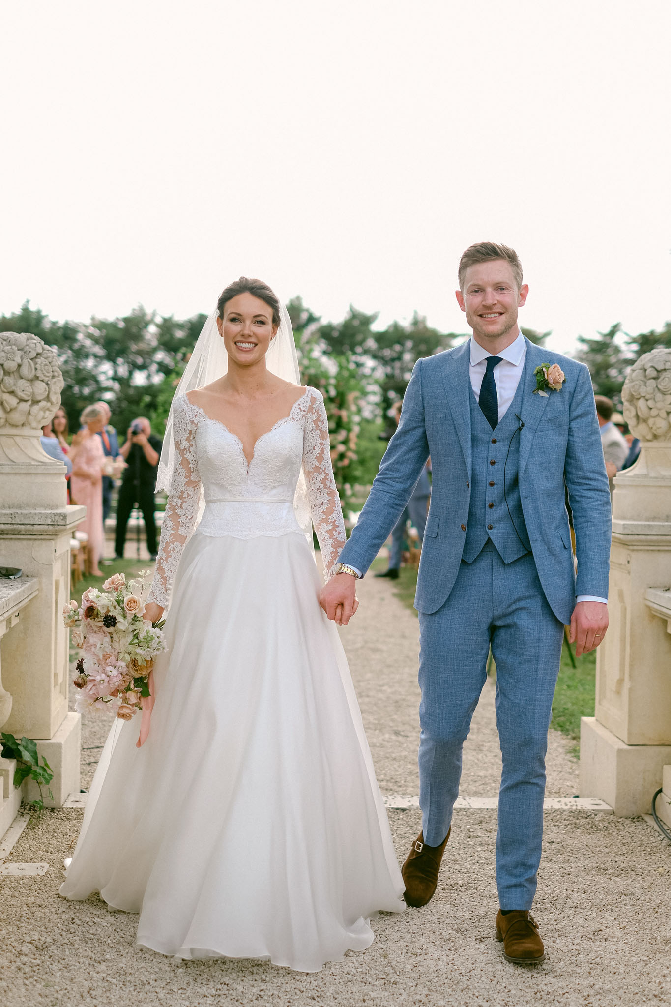 Bride with blush anemone bouquet and groom in blue checked suit recessioning past stone balustrade