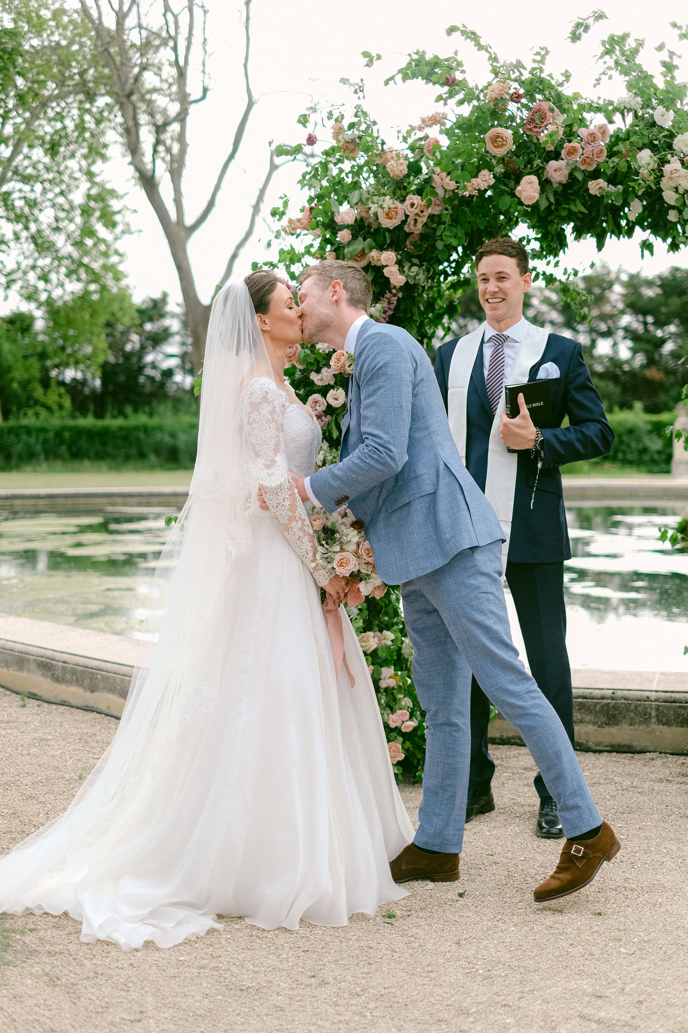 The first kiss moment of an outdoor wedding ceremony, captured in a full-length portrait shot. The bride wears a long-sleeve lace bodice ball gown with a cathedral-length veil and holds a bouquet of blush, peach, and dusty rose garden roses with trailing greenery. The groom wears a light blue suit with brown suede monk-strap shoes. Behind them, an officiant in a navy suit holds a Holy Bible and smiles broadly. The ceremony takes place beside a formal rectangular reflecting pool, in front of a large floral arch densely covered in blush, peach, ivory, and mauve garden roses with lush green foliage. The overall decor palette is soft blush and peach with a classic garden aesthetic.