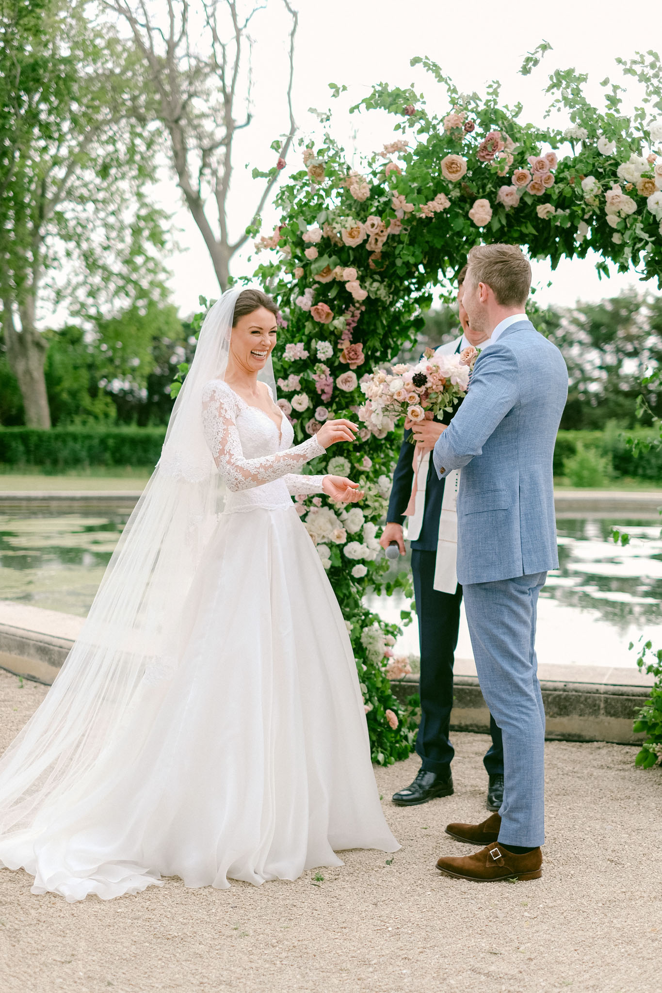An outdoor wedding ceremony ring exchange moment, with the bride laughing as she extends her hand toward the groom. The ceremony takes place beside a formal rectangular reflecting pool, with a large floral arch behind the couple featuring blush, dusty pink, and cream garden roses, anemones, and lush greenery. The bride wears a long-sleeve lace-bodice ballgown with a full tulle skirt and a cathedral-length veil; the groom is dressed in a light blue suit with brown monk-strap shoes. A third figure, likely the officiant, stands between and behind them in a navy suit, holding a microphone and the bride's bouquet of blush and cream garden roses. The shot is a medium full-length portrait taken at ground level.