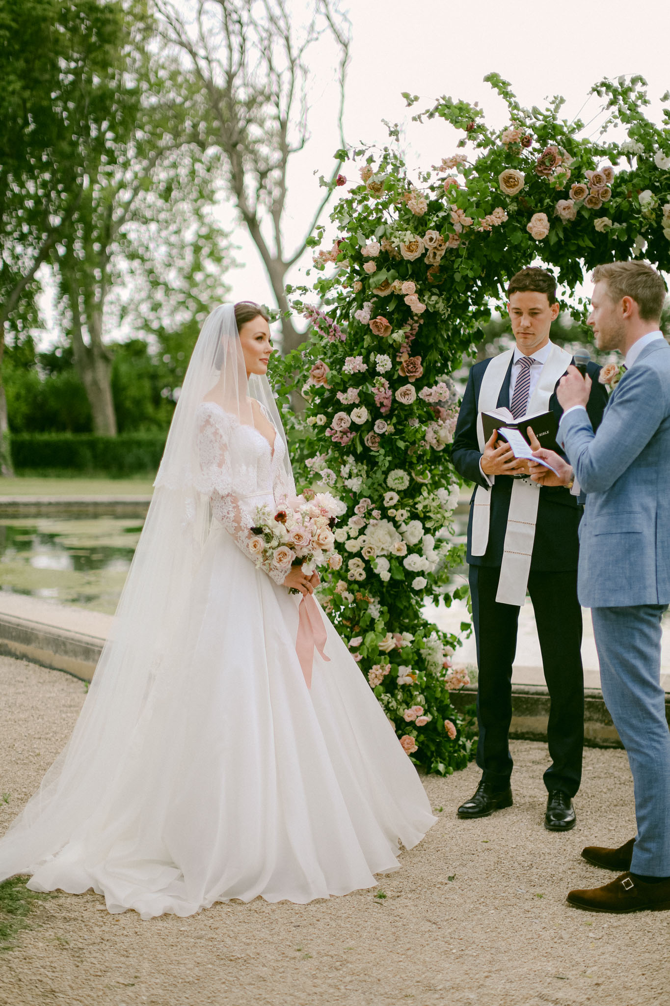 Ceremony beside reflecting pool with circular blush and dusty rose arch, bride in lace gown and cathedral veil