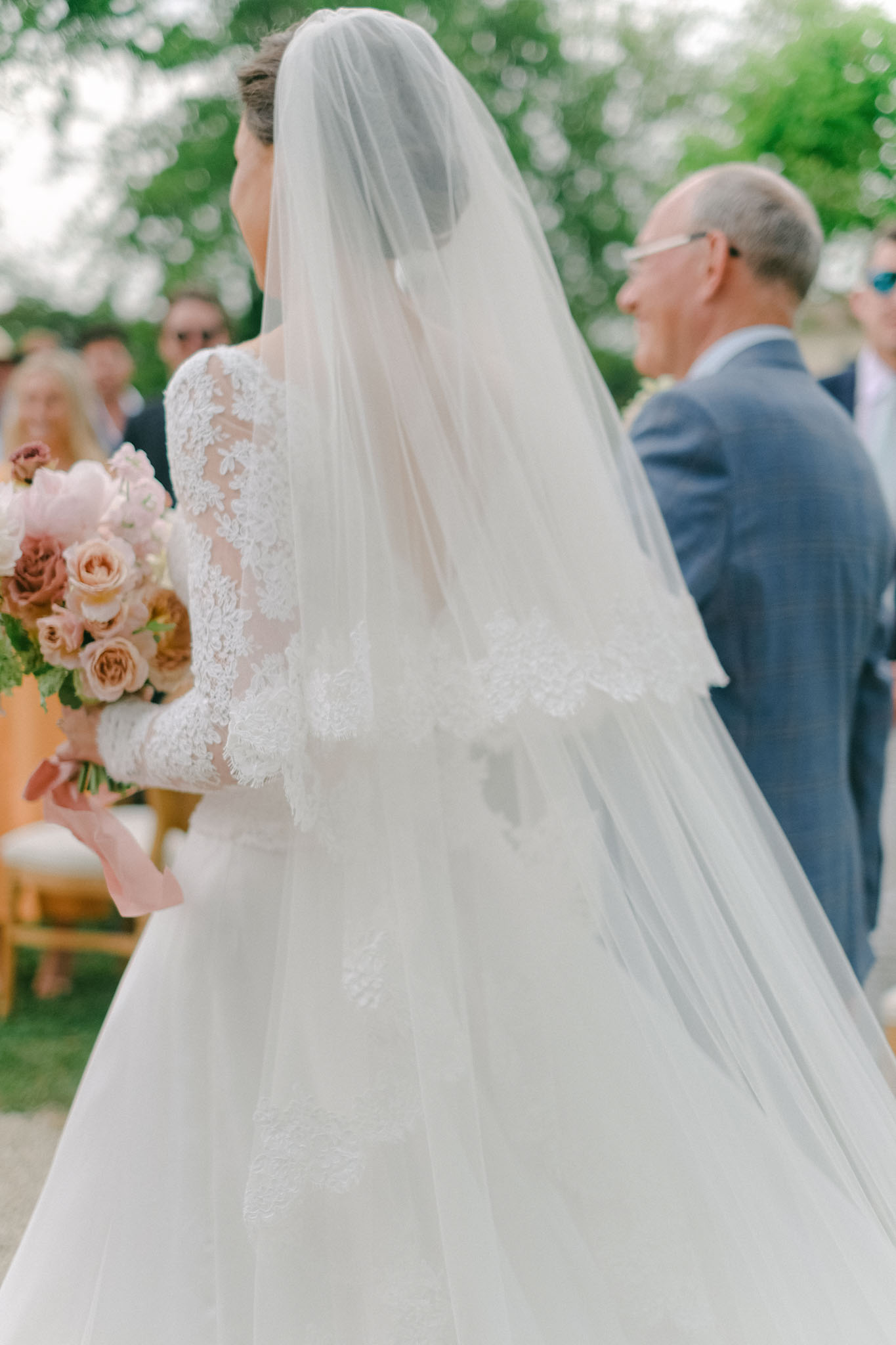 Bride from behind with lace-edged cathedral veil and blush peony bouquet walking down aisle with escort