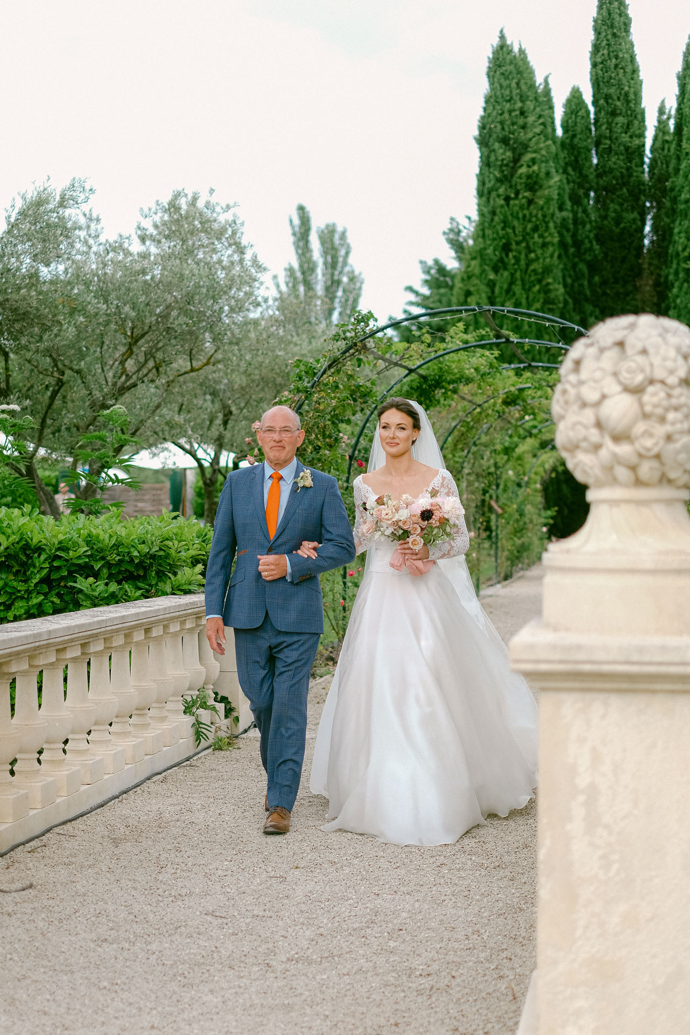 Bride in lace-sleeve ballgown walking with father past stone balustrade and cypress trees