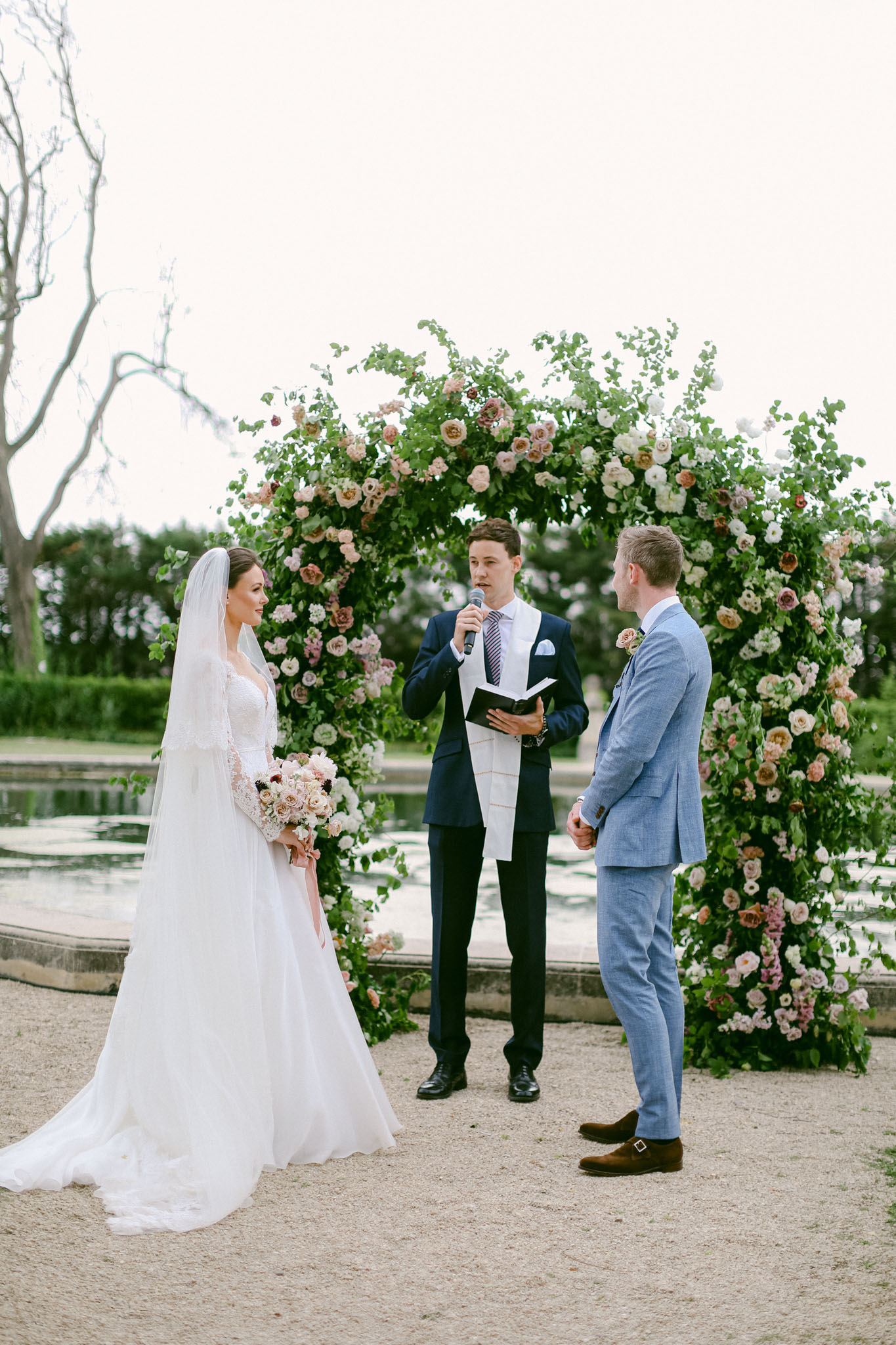 An outdoor wedding ceremony taking place on a gravel terrace in front of a formal reflecting pool or ornamental water feature. The bride wears a long-sleeved lace bodice gown with a full tulle skirt and a cathedral-length veil, holding a bouquet of blush, ivory, and dusty rose garden roses and ranunculus. The groom wears a light blue suit with brown monk-strap shoes and a small floral buttonhole. An officiant in a navy suit with a striped tie and white ceremonial stole speaks into a handheld microphone while holding an open book. The three stand beneath a large circular floral arch densely covered in green foliage and layered with blush, ivory, caramel, and dusty pink garden roses and ranunculus. The ceremony style is classic and garden-inspired. Medium full-length portrait shot capturing all three figures from the front.