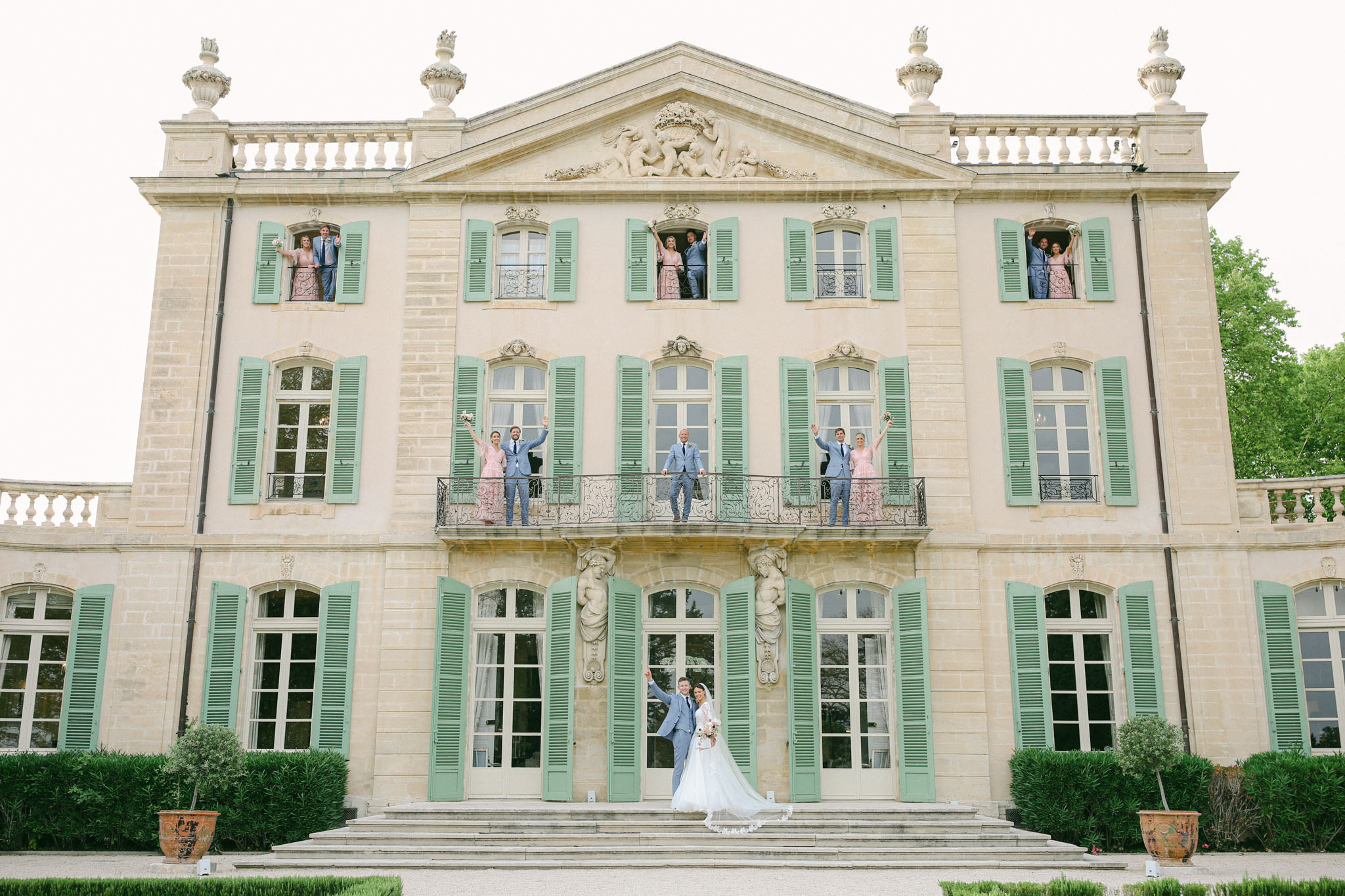 A wide-shot bridal party portrait taken outdoors in front of a three-story French château with pale pink stone facade, sage green shutters, ornate carved stonework, and a wrought iron balcony. The bride and groom stand at the base of the front steps at ground level — the bride in a long white ball gown with a cathedral veil and the groom in a light blue suit — both with arms raised in celebration. Three pairs of bridesmaids and groomsmen are positioned across the second-floor balcony, each pair consisting of a woman in a blush pink lace dress and a man in a light blue suit, arms raised. Three additional pairs are posed leaning out of the upper-floor windows in the same attire, creating a symmetrical, playful composition across all three levels of the building. The styling palette is cohesive with blush pink and light blue repeated throughout the bridal party. Terracotta urns with topiaries flank the entrance steps. Potential venue feature image.