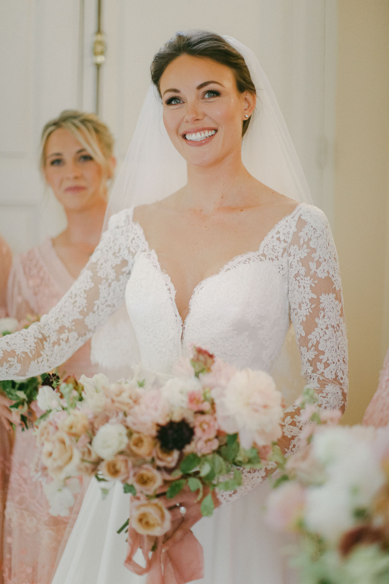 Bride in white lace gown with blush rose and mauve anemone bouquet flanked by bridesmaids indoors