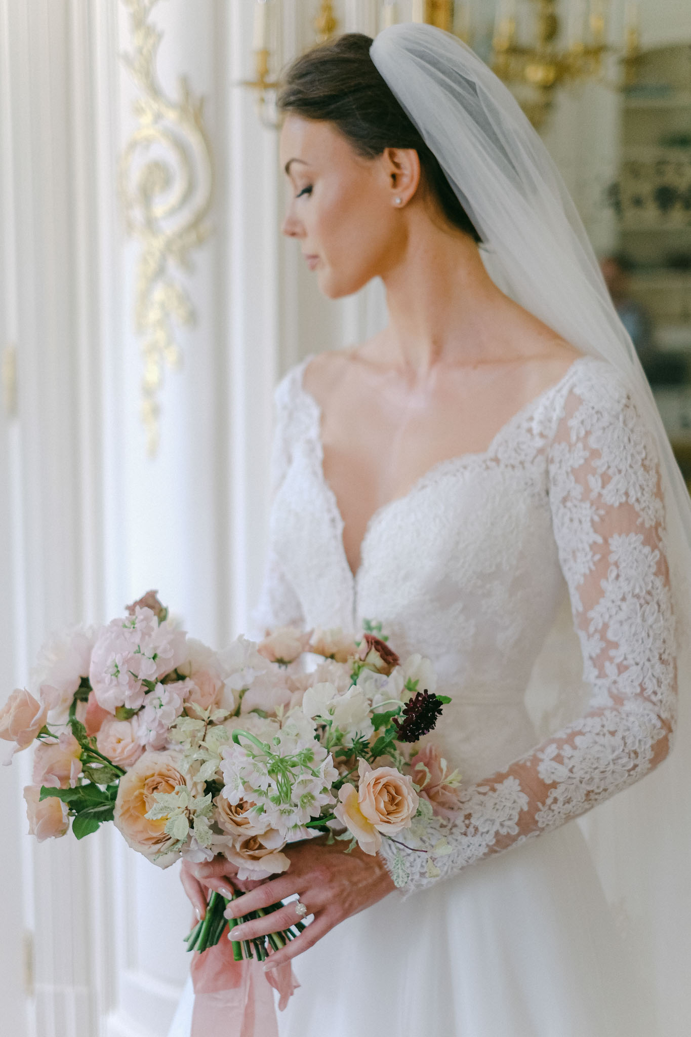 Bride in lace gown with cathedral veil holding blush sweet pea and peach rose bouquet in gilded salon