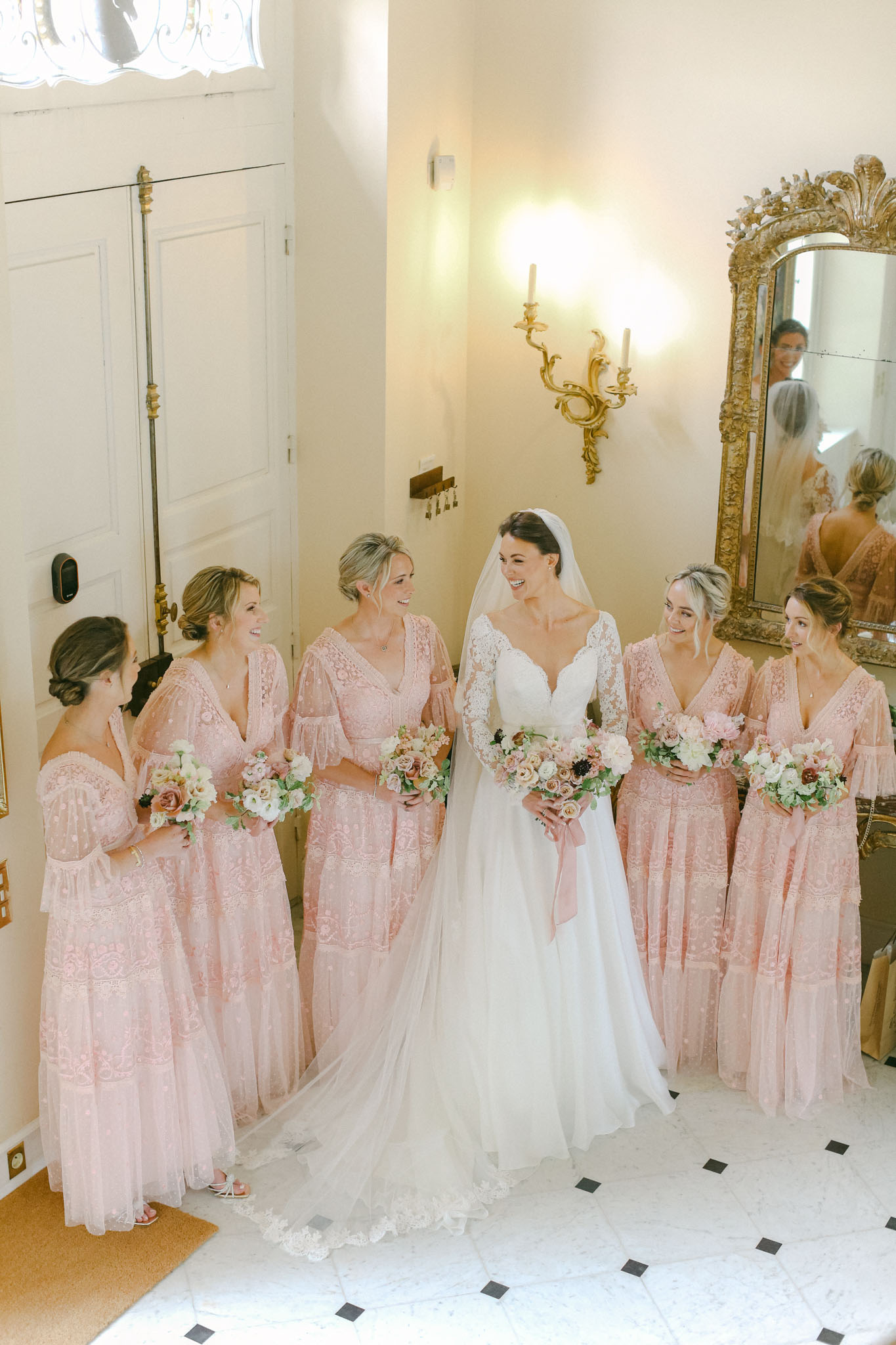 Bride and four bridesmaids in blush lace dresses laughing on marble floor beside gilt mirror