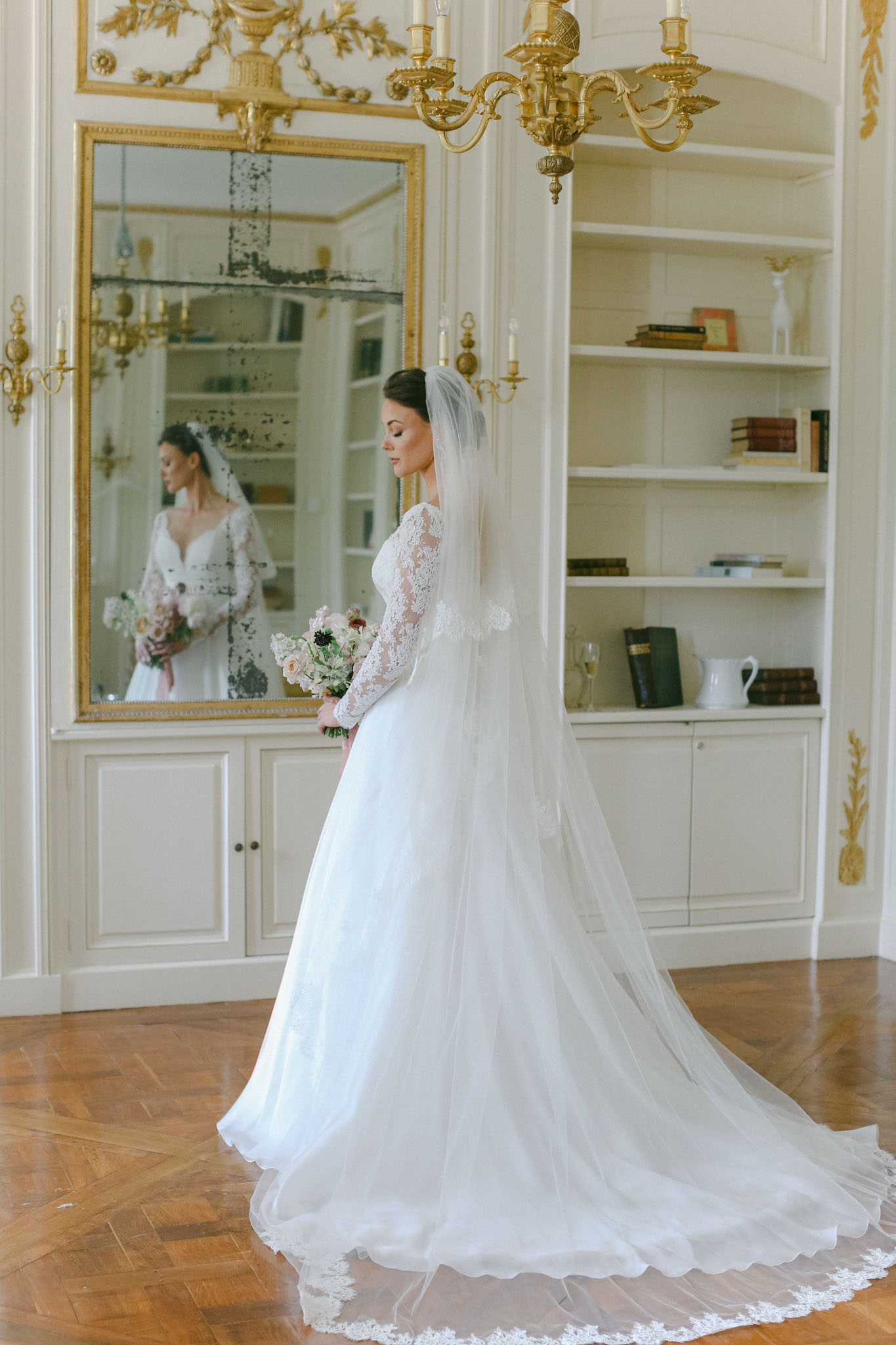 Bride in white lace ball gown with cathedral veil standing in gilded French interior with mirror reflection