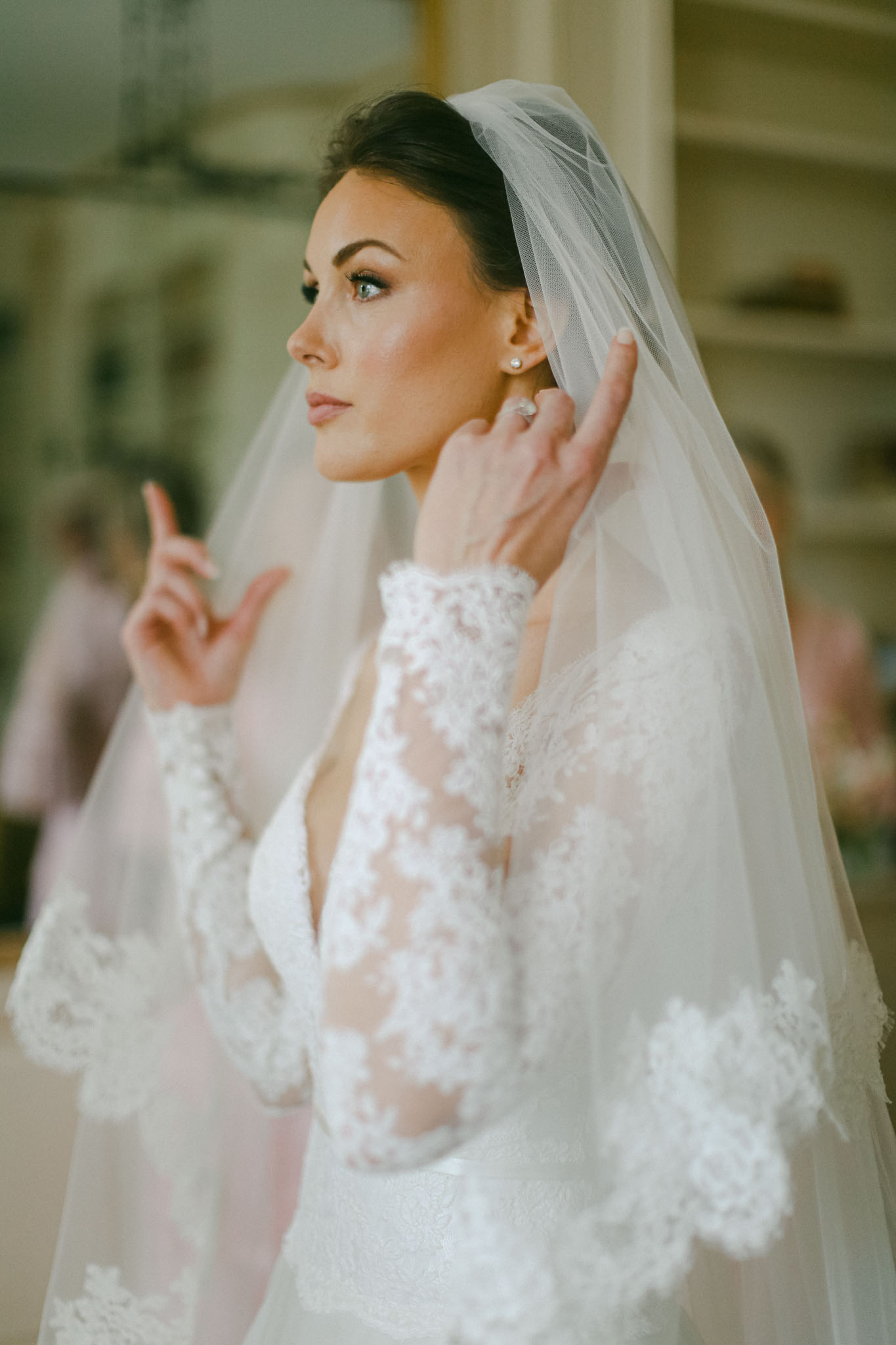 Bride adjusts pearl drop earring wearing lace applique gown and cathedral veil with low updo close-up