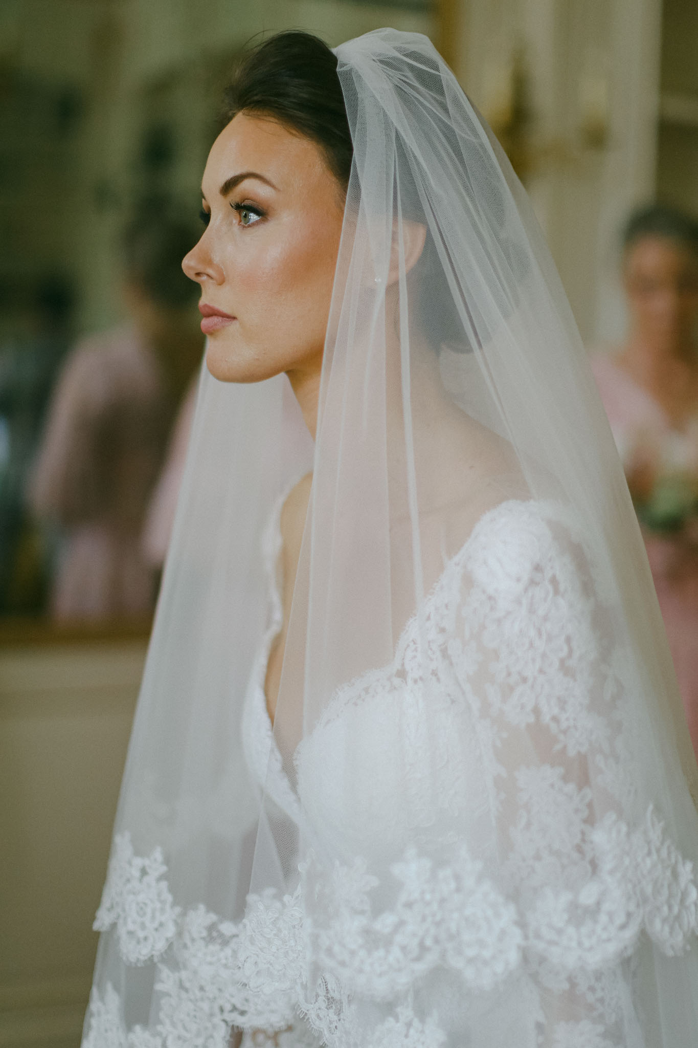 Side profile bride in long-sleeve lace gown with tulle veil and updo before gilded wall panelling