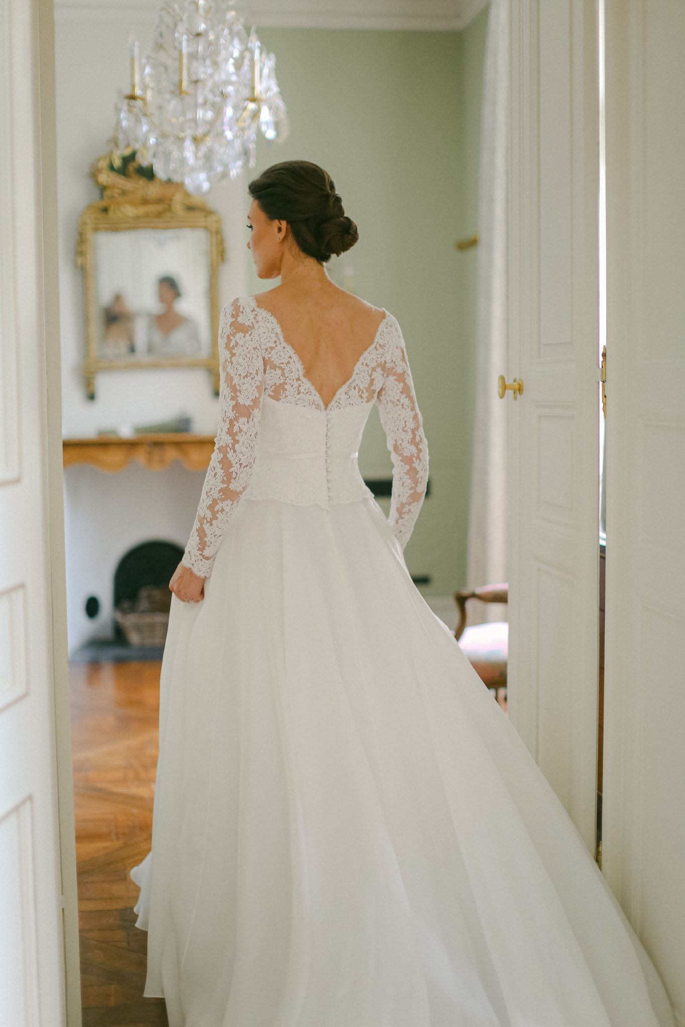 A bridal portrait taken indoors, showing the bride from behind as she stands in a doorway. She wears an ivory gown with long lace sleeves, a deep V-back with lace detail, a fitted lace bodice, and a full flowing skirt with a train. Her dark hair is styled in a low updo. The room behind her features a crystal chandelier, a gilded gold ornate mirror above a carved stone fireplace, herringbone parquet flooring, and pale sage-green walls, consistent with a classic French château interior. A photographer's reflection is faintly visible in the mirror. The composition is a three-quarter portrait shot from behind, framed by the white double doors with brass hardware.