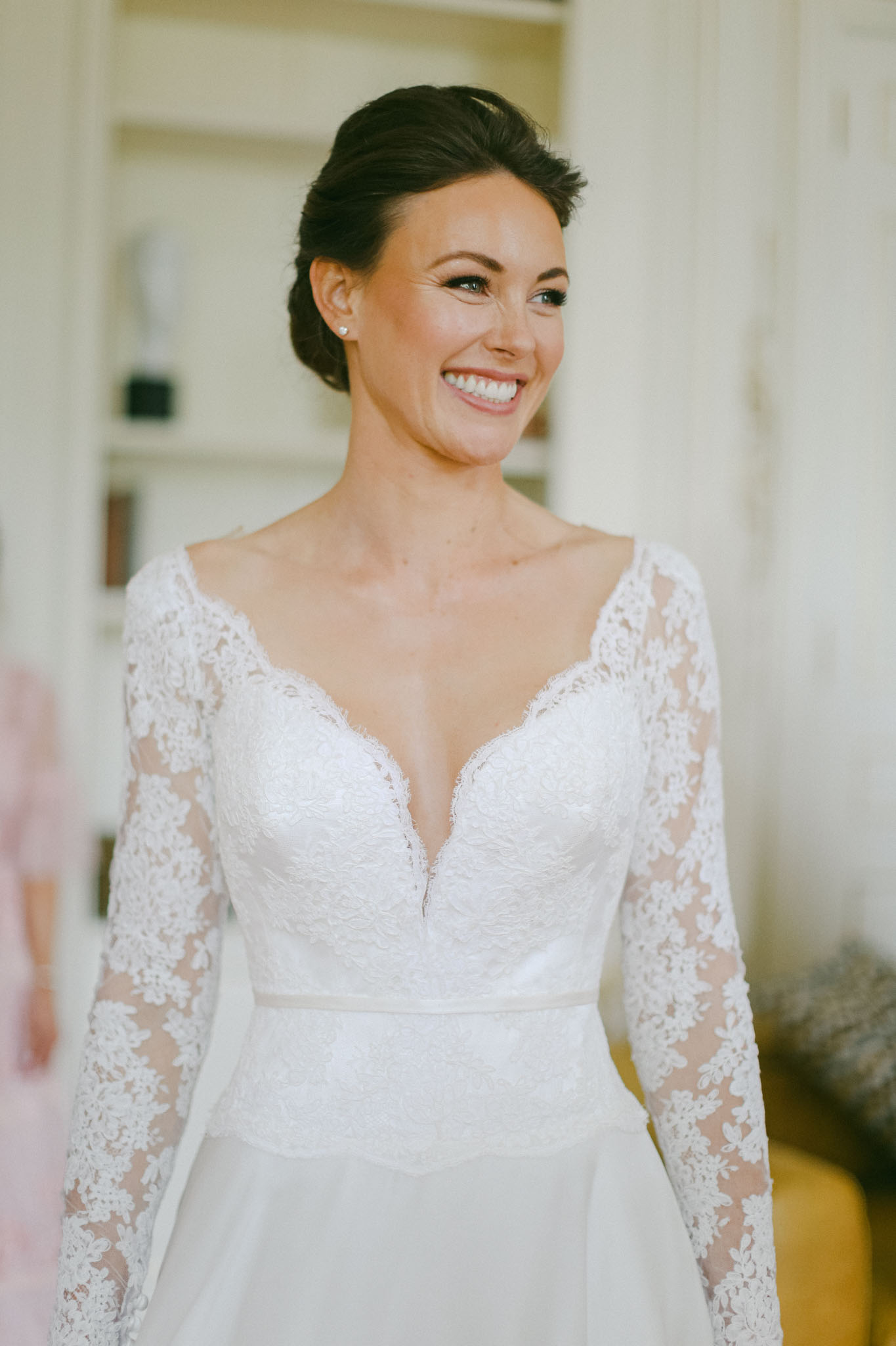 A close-up portrait of a bride during the getting-ready phase, photographed indoors in a light-filled room with neutral white walls and a mirror visible in the background. She is wearing a white lace wedding gown with long sheer lace sleeves, a deep V-neckline with scalloped lace detailing, and a satin skirt with a thin belt at the waist. Her dark brown hair is styled in a low updo, and she wears small stud earrings. Her makeup includes defined brows, smoky eye liner, and a natural pink lip. She is smiling and looking slightly off-camera to one side.