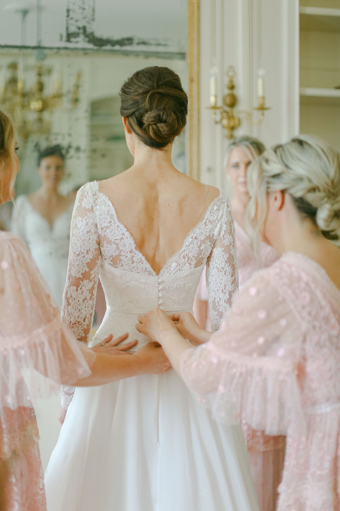 A getting-ready scene captured indoors in what appears to be a classically decorated room with white panelling and gold wall sconces fitted with candles. The bride stands with her back to the camera while two bridesmaids in blush pink sequined robes fasten the back of her wedding dress. The gown features three-quarter-length sheer lace sleeves, a deep V-back neckline, floral lace bodice with covered buttons running down the back, and a flowing ivory skirt. The bride's dark hair is styled in a twisted chignon updo. A large gilt-framed mirror in the background reflects the smiling face of one bridesmaid and the front of the bride's dress. The composition is a close-up portrait shot focused on the back of the dress and the hands of the bridesmaids assisting with the fastening.