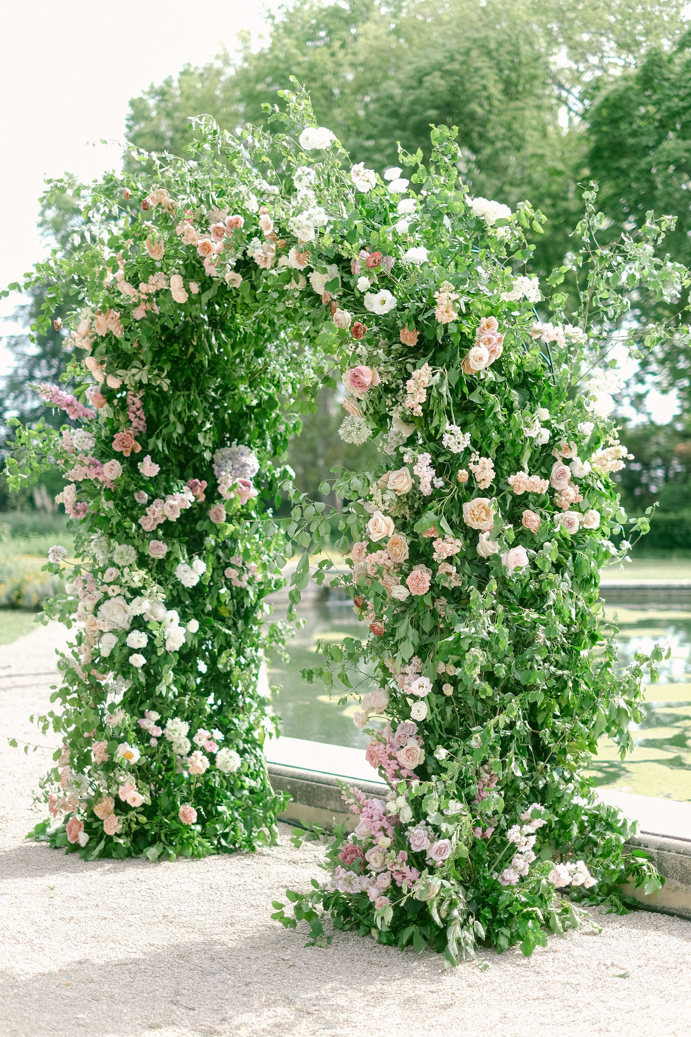 Lush floral arch installation with blush, peach, and mauve garden roses beside a reflecting pool