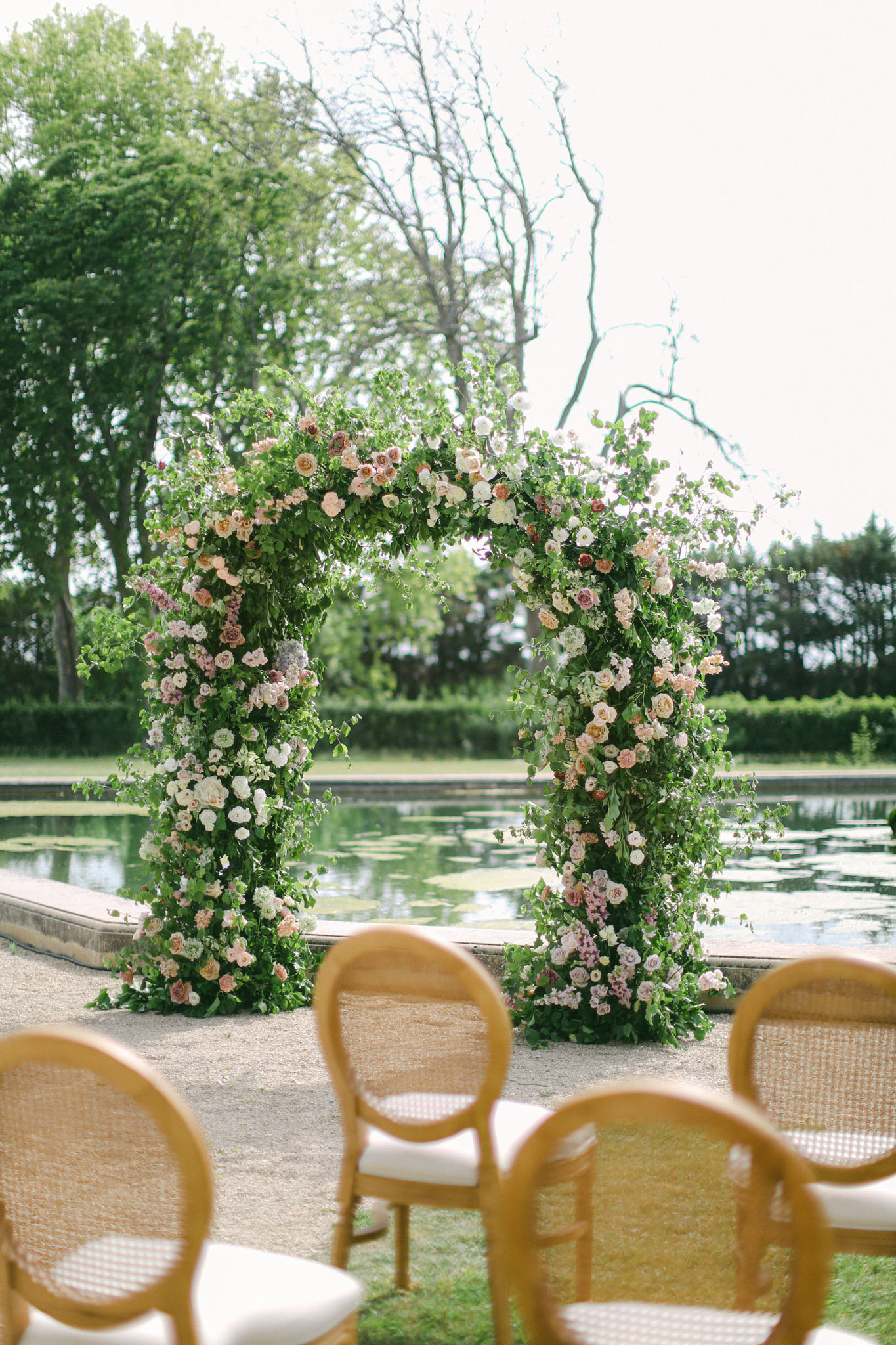 Outdoor ceremony setup with dense floral arch of garden roses at ornamental pond edge, cane-back chairs in rows