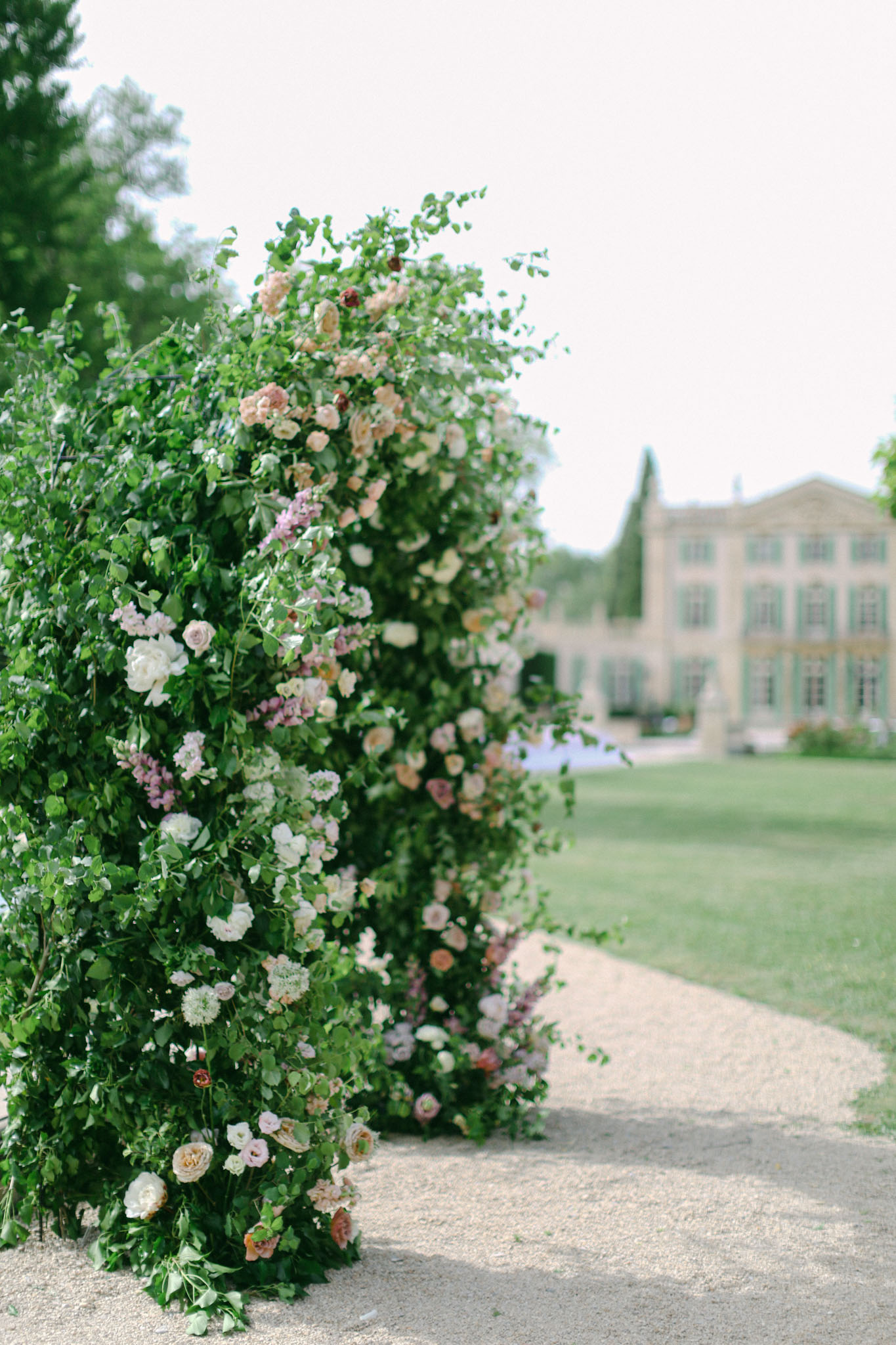 Floral arch with ivory roses, blush and mauve roses, peach ranunculus, and wisteria on gravel path near chateau