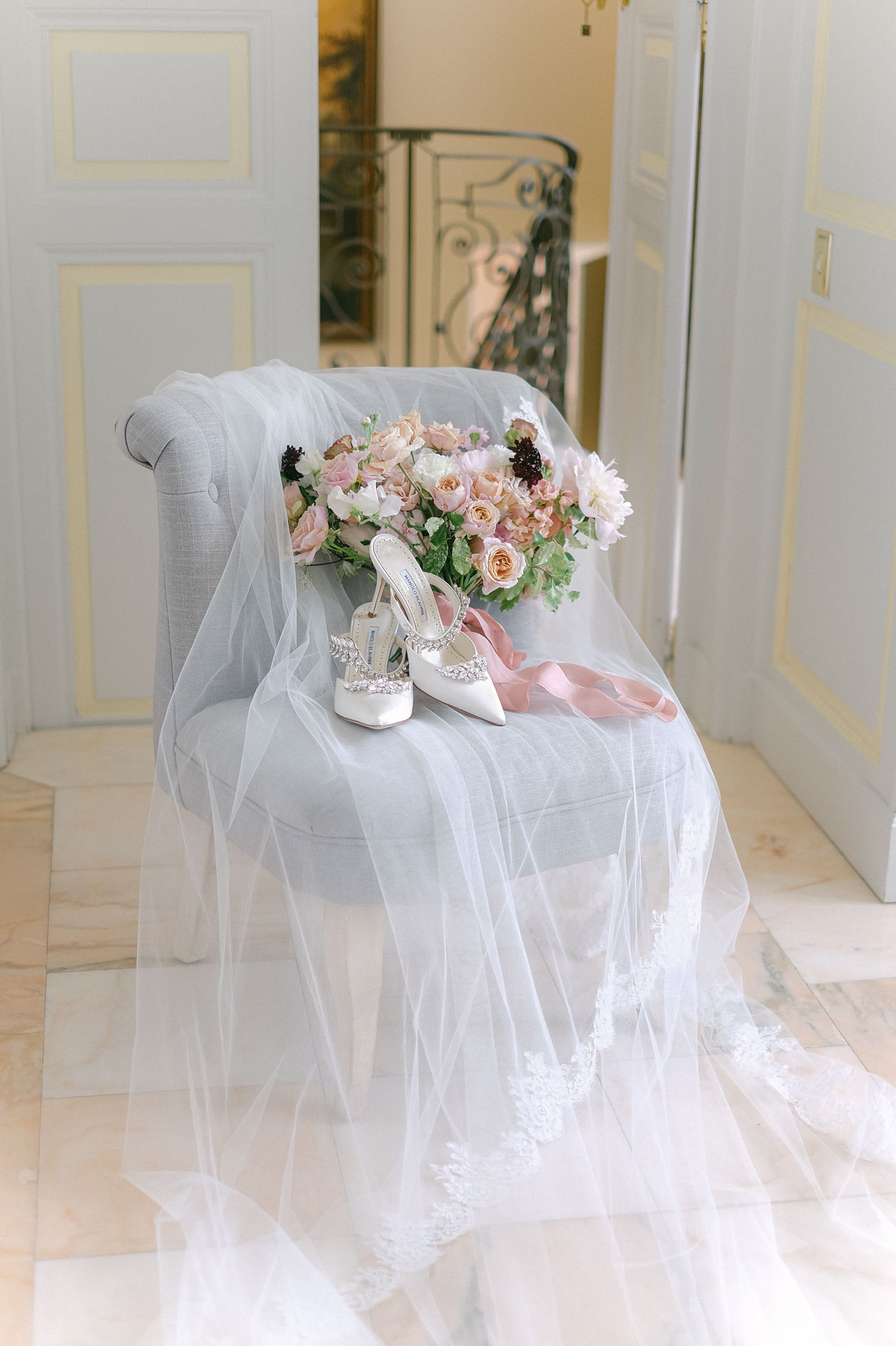 A bridal flat lay styled on a light grey upholstered armchair inside what appears to be a château or formal interior, identifiable by white panelled walls, marble tile flooring, and a wrought iron staircase visible through an open doorway in the background. A tulle veil with lace appliqué trim drapes over the chair and spills onto the floor, with a pair of white satin Manolo Blahnik pointed-toe heels embellished with crystal buckle detailing placed at the center. A bridal bouquet composed of blush pink roses, peach garden roses, ivory blooms, dark burgundy accents, and green foliage rests against the chair back, with a dusty pink satin ribbon trailing across the seat. The composition is a medium portrait-style detail shot with soft, bright natural light and a classic, romantic styling aesthetic.
