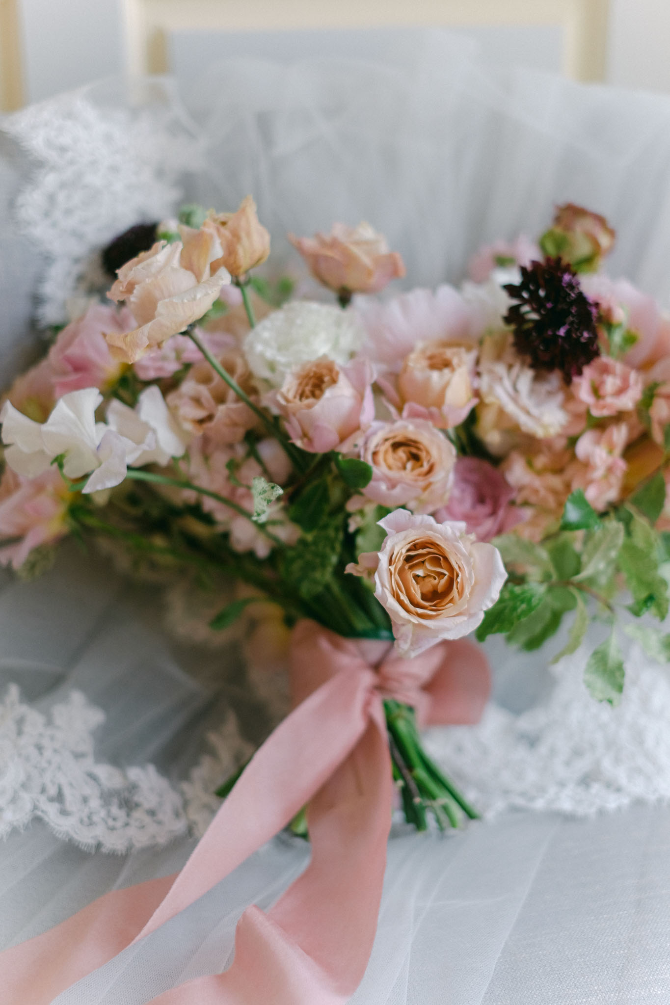 A close-up detail shot of a bridal bouquet resting on a tulle veil with a lace trim edge. The bouquet is composed of peach garden roses, blush pink sweet peas, white ranunculus, pale pink spray roses, and a deep burgundy scabiosa, with green foliage throughout. The stems are bound with a long dusty pink satin ribbon tied in a trailing bow. The overall floral palette is soft and romantic, mixing warm peach and blush pink tones with dark burgundy accents. The styling is loose and garden-inspired rather than tightly structured.